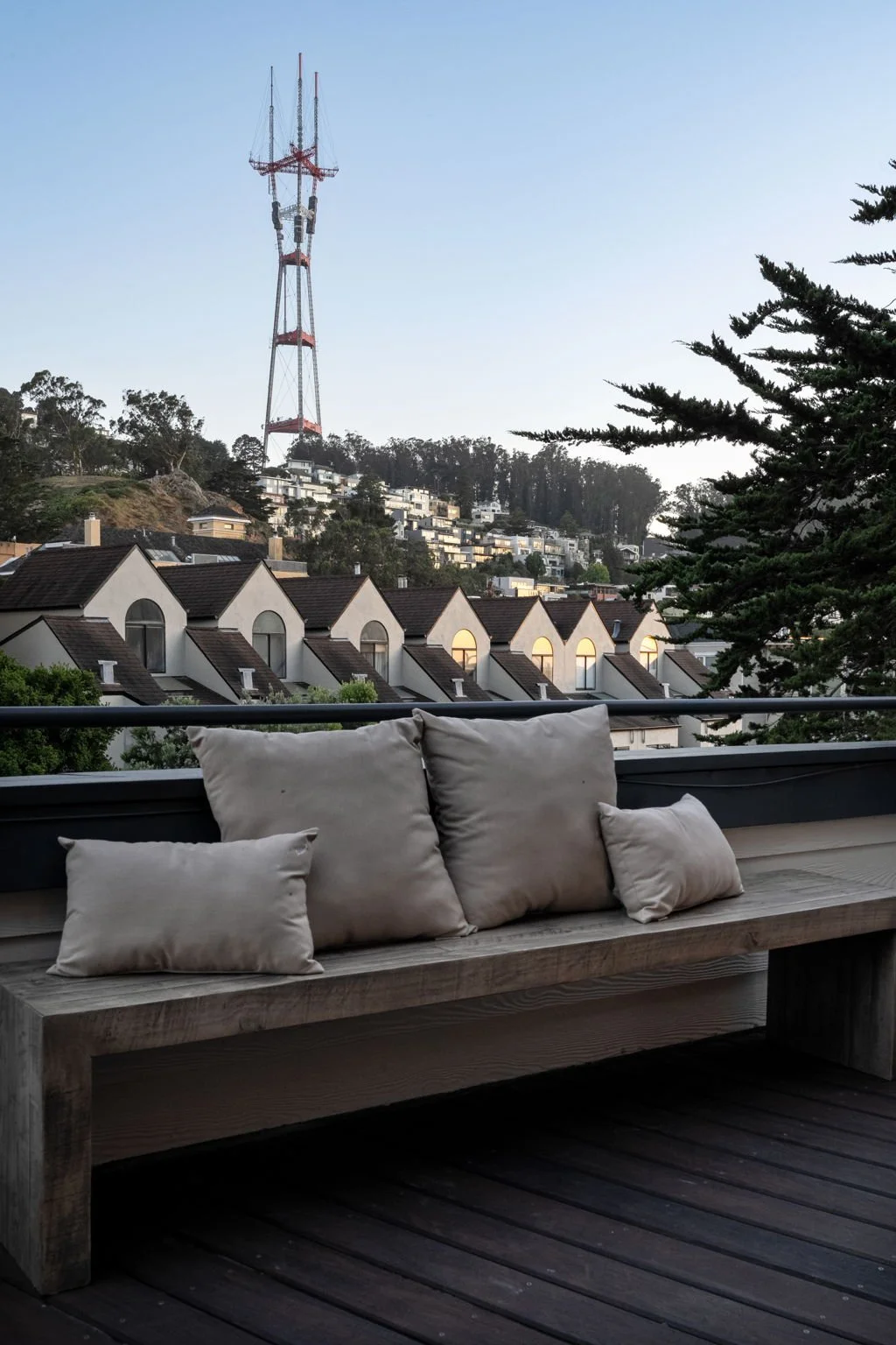 A balcony with beige pillows on a wooden bench overlooking a residential area with houses and a hillside with a radio tower.