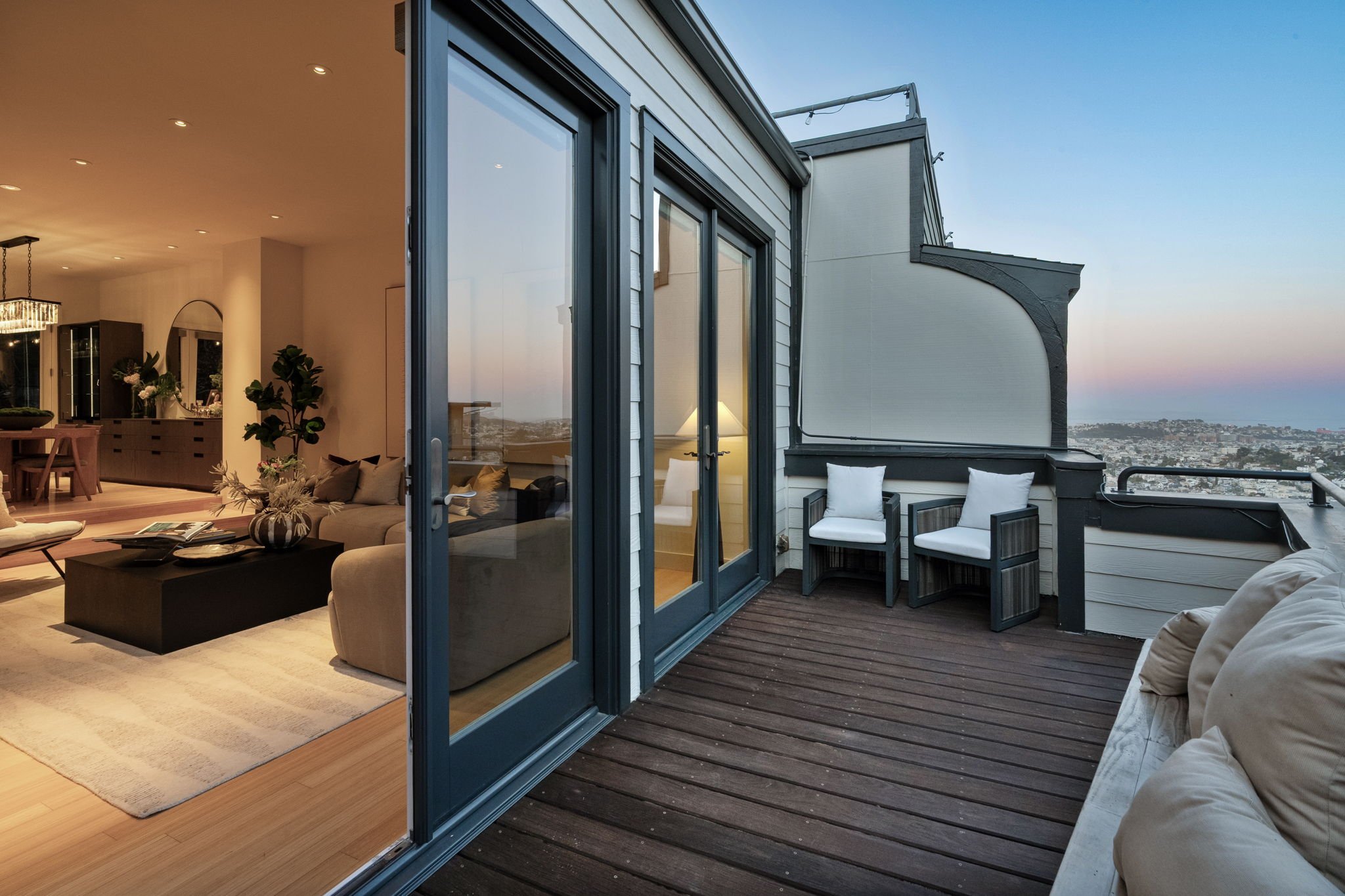 Living room with beige sofas, a black coffee table with magazines, and a dining area with a chandelier, as seen from a balcony with outdoor chairs and a view of a cityscape and ocean during dusk.
