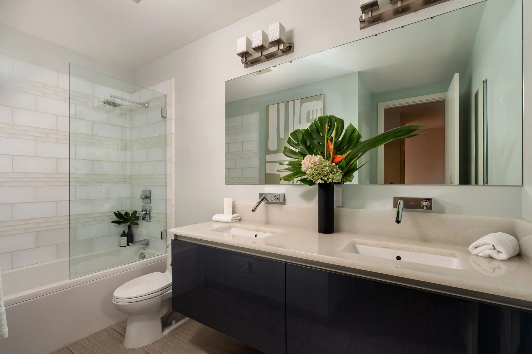 Modern bathroom with a large mirror, a black vanity with a white countertop, a flower arrangement in a black vase, a white towel, a glass-enclosed shower with white subway tiles, a built-in niche, a rainfall showerhead, and a white toilet.