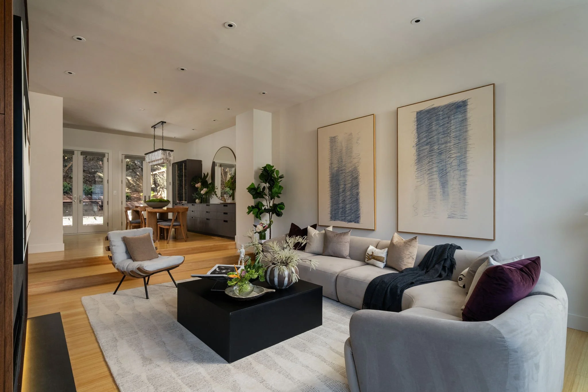 Living room with a cream sofa, pillows, coffee table, modern chairs, artwork, and plants