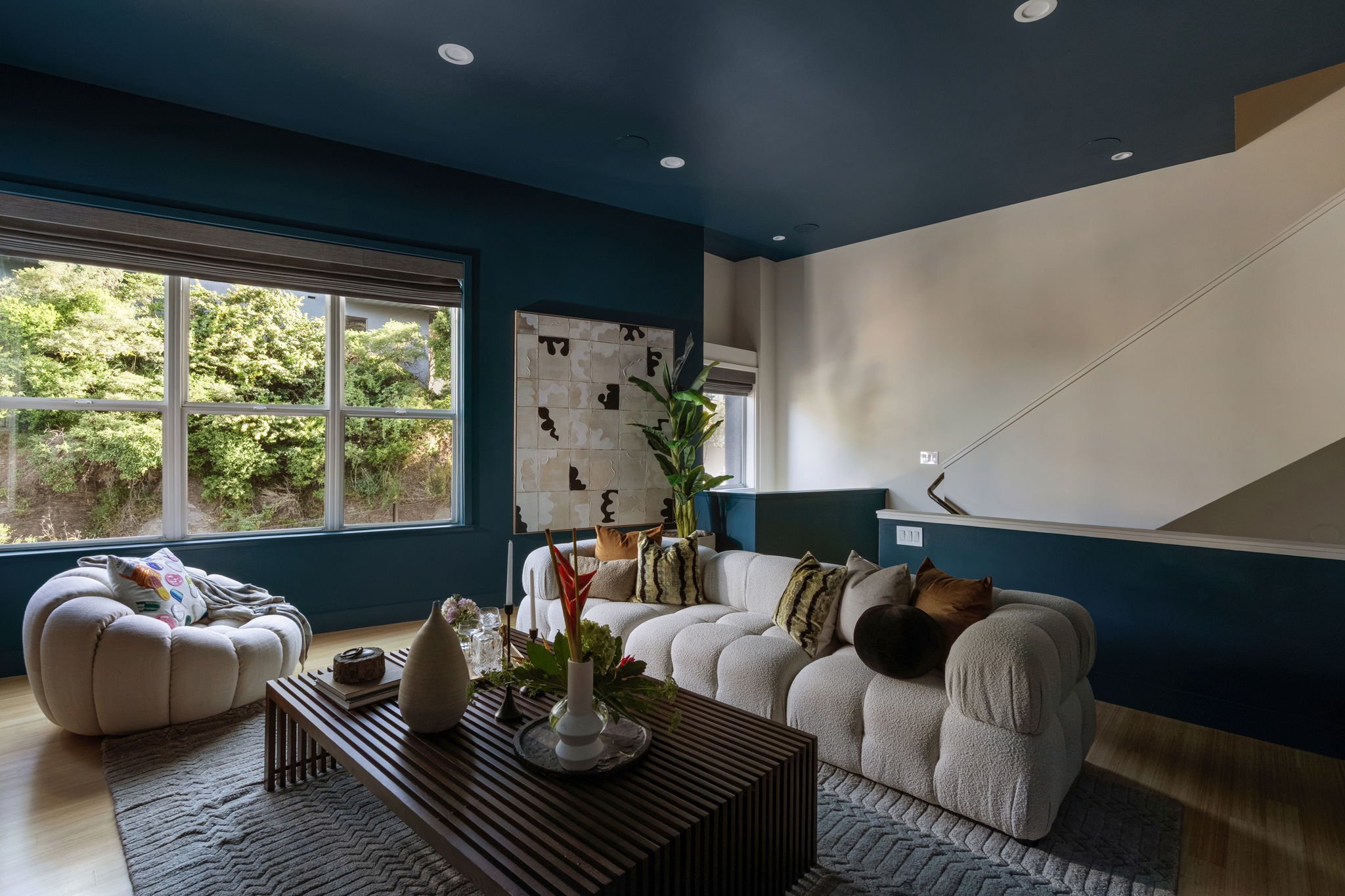 A living room with large windows showing green trees outside. The room has blue and beige walls, a beige plush sofa with textured and patterned cushions, a wooden coffee table with decorative vases and candles, a patterned area rug, and a potted plan