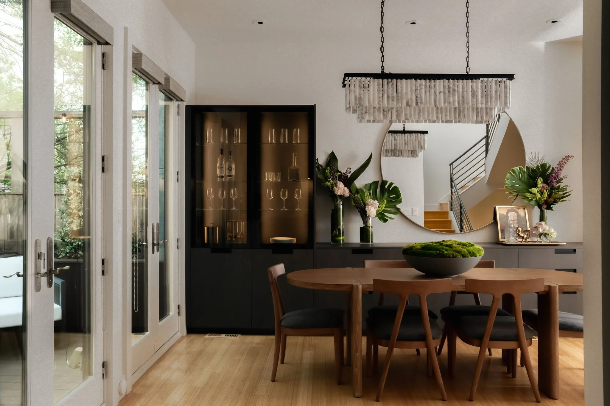 Interior of a modern dining room with wooden table and chairs, glass doors, a black cabinet with glassware, decorative flowers, a large round mirror, and a staircase in the background.