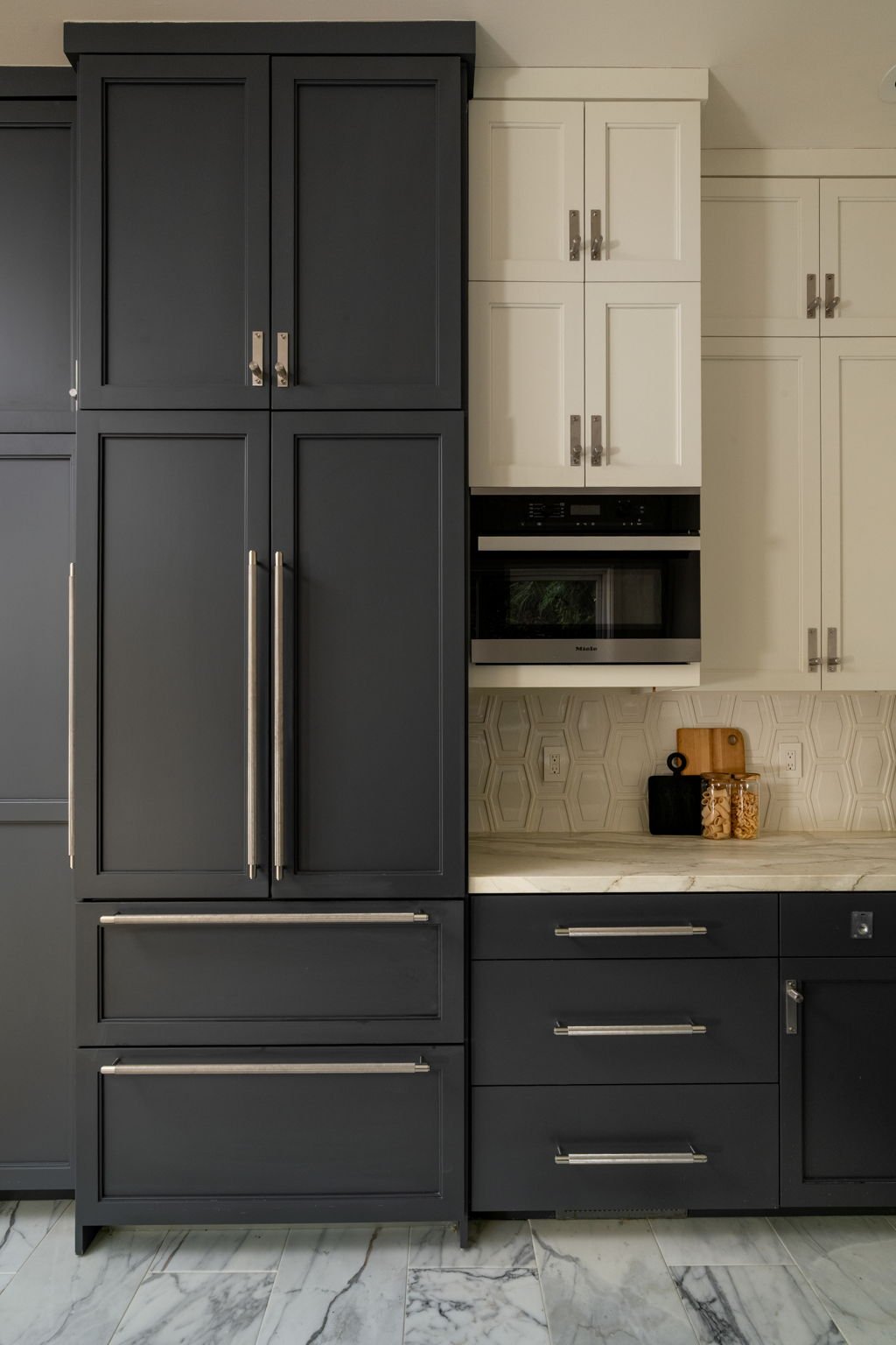 Kitchen cabinets with black and cream drawers, countertop with cutting board and jars, microwave in a beige kitchen.