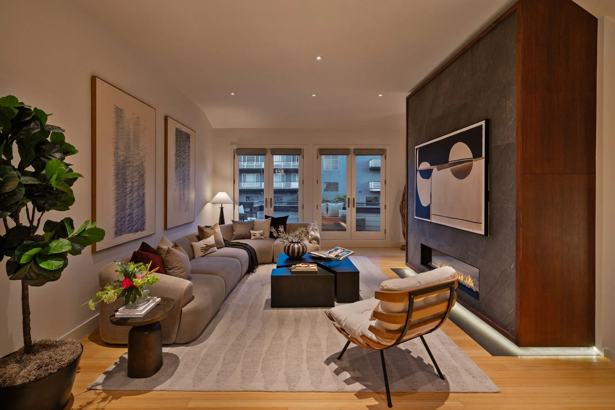 A modern living room with a large beige sectional sofa, several throw pillows, a black coffee table, and a wooden armchair with a cushion. There are abstract artwork pieces on the wall, a potted plant, a floor lamp, and large windows with views of a 