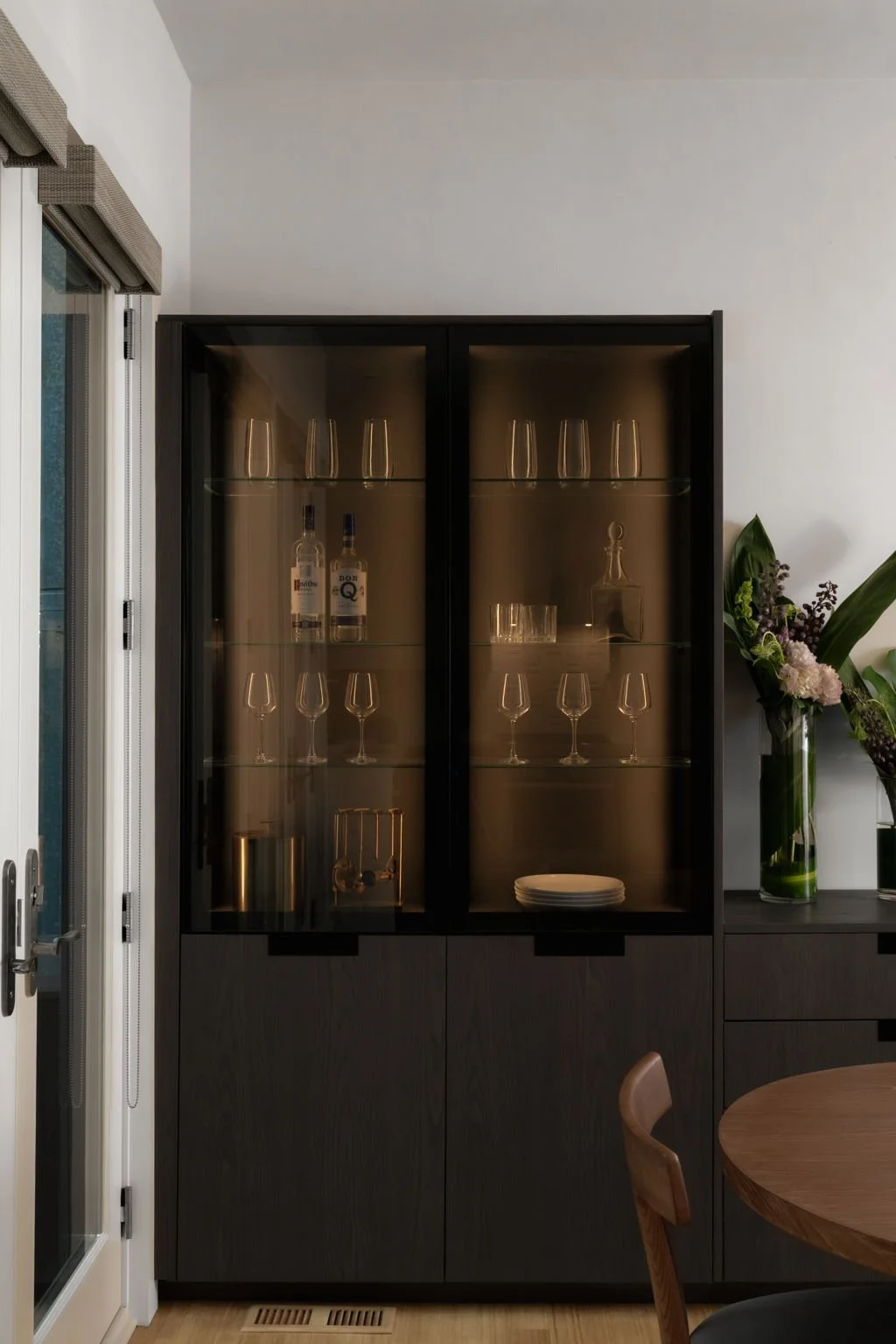 A black display cabinet with glass doors showing glassware and bottles, located next to a door with window and a flower arrangement on the side.