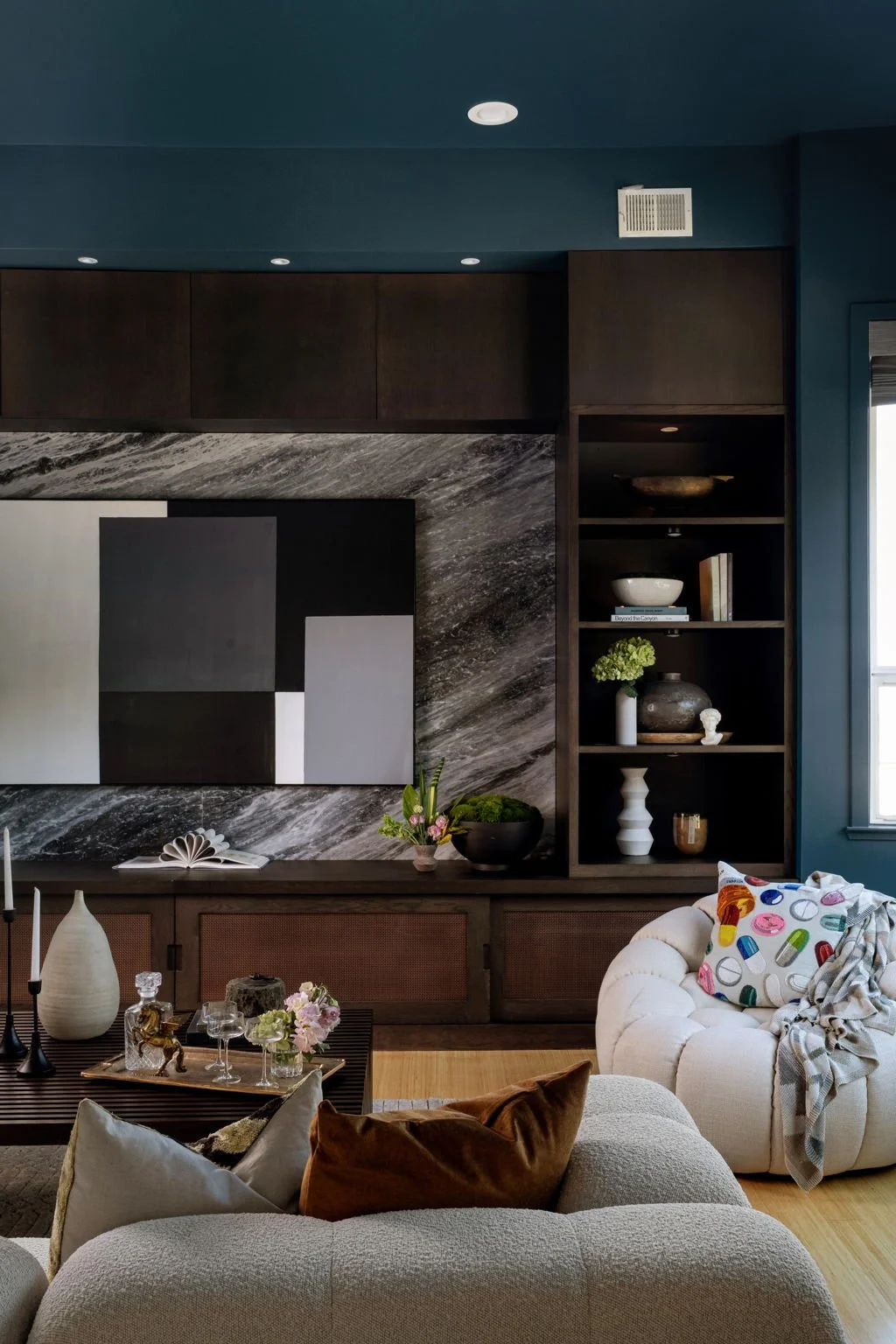 A modern living room with a dark built-in bookshelf displaying decorative items such as vases, bowls, and books, a dark geometric abstract artwork, a gray and beige sofa with pillows, and a coffee table with candles, flowers, and glasses, all under a