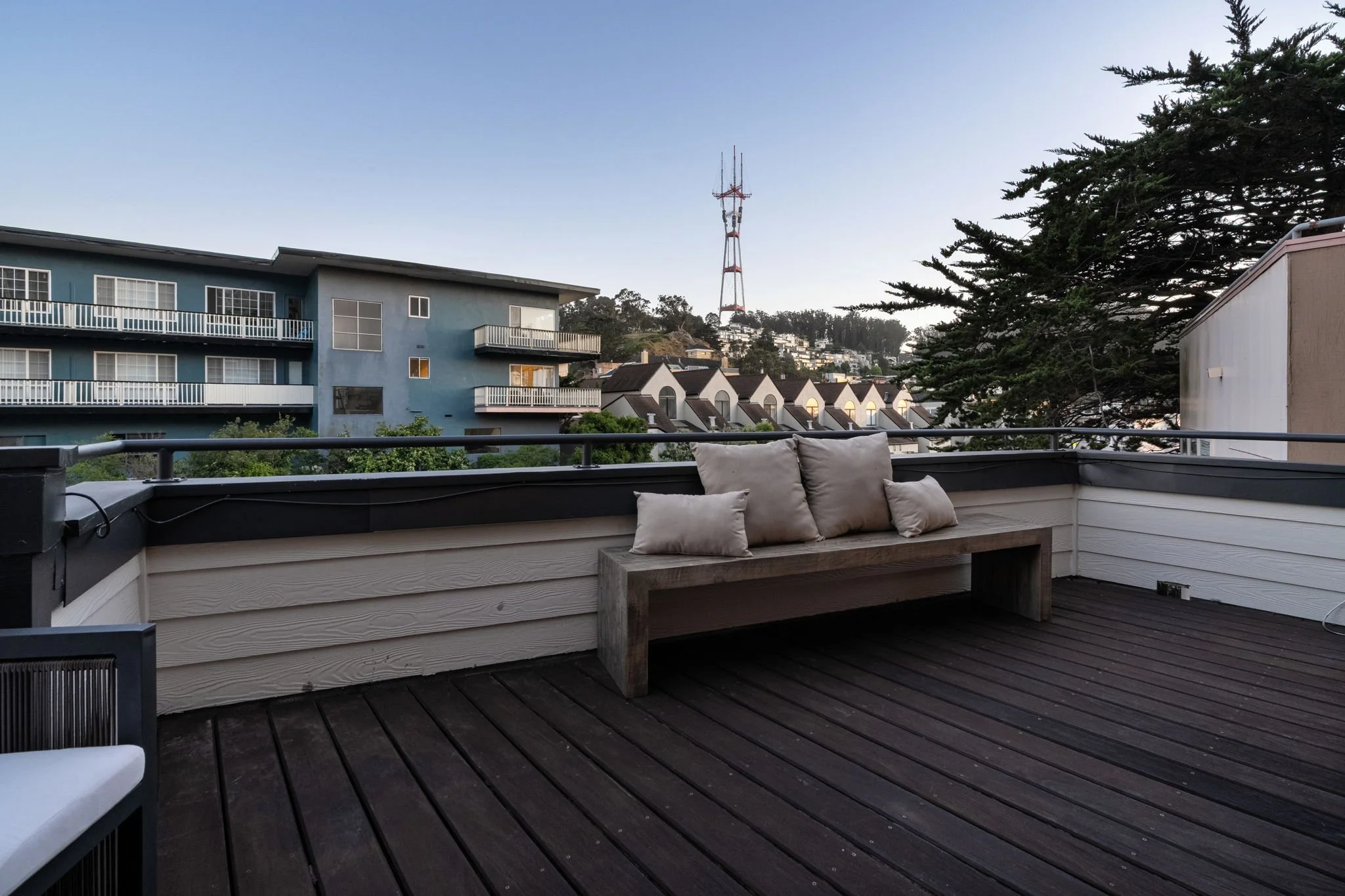 Balcony with wooden flooring and cushions on a built-in bench, overlooking neighboring buildings and a large communication tower on a hill in the distance, under a clear sky.