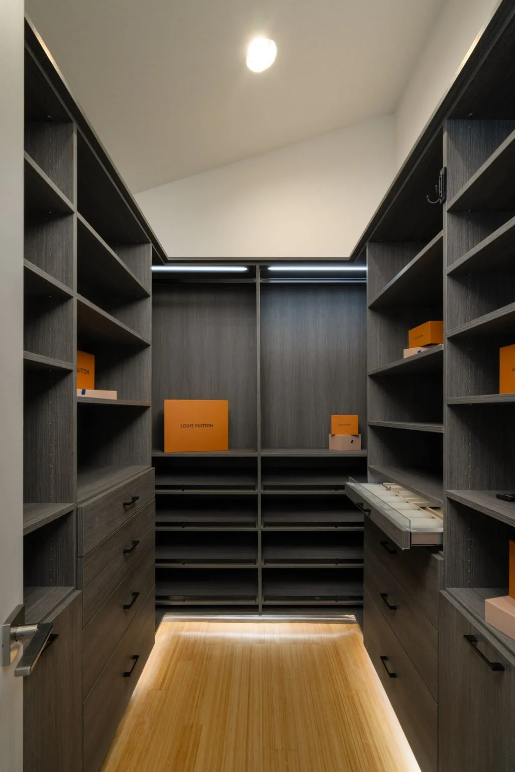 A walk-in closet with dark gray wood shelving and drawers, containing orange Louis Vuitton boxes, under ceiling lighting, with a wooden floor.