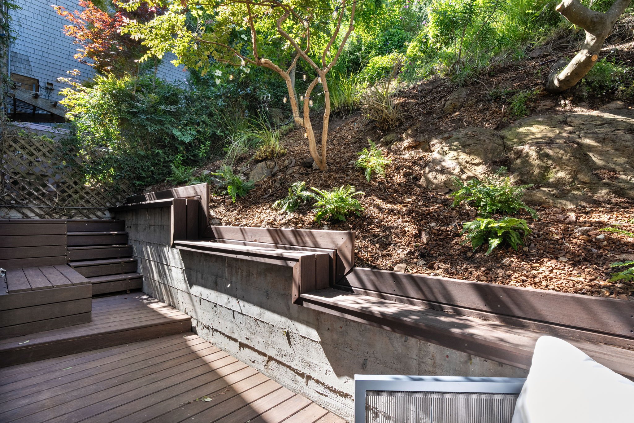 A backyard with wooden stairs leading up to a mulched garden slope filled with various green plants, bushes, and trees, some with colorful leaves, and a gray wood-paneled house wall in the background.