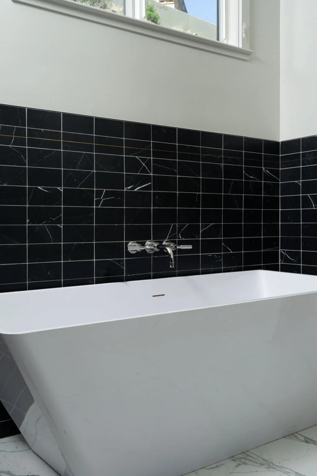 A modern freestanding bathtub with a black marble tiled wall, a silver faucet, near a window with a view of greenery.