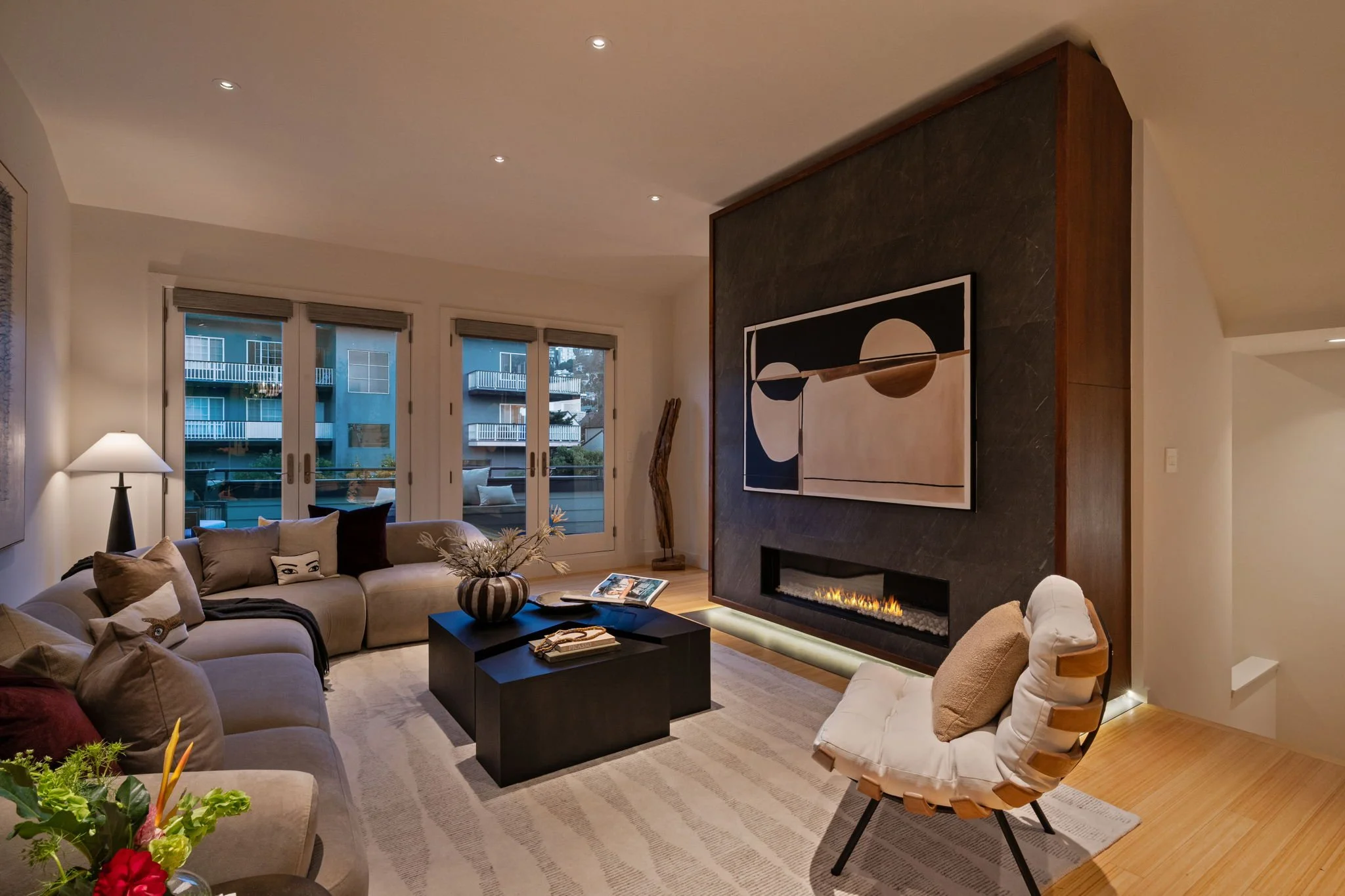 Modern living room with a large beige sectional sofa, black coffee table, and a white armchair with wooden armrests. A fireplace with a black stone surround and a bookshelf-like fire feature is on one side. There are three large sliding glass doors w