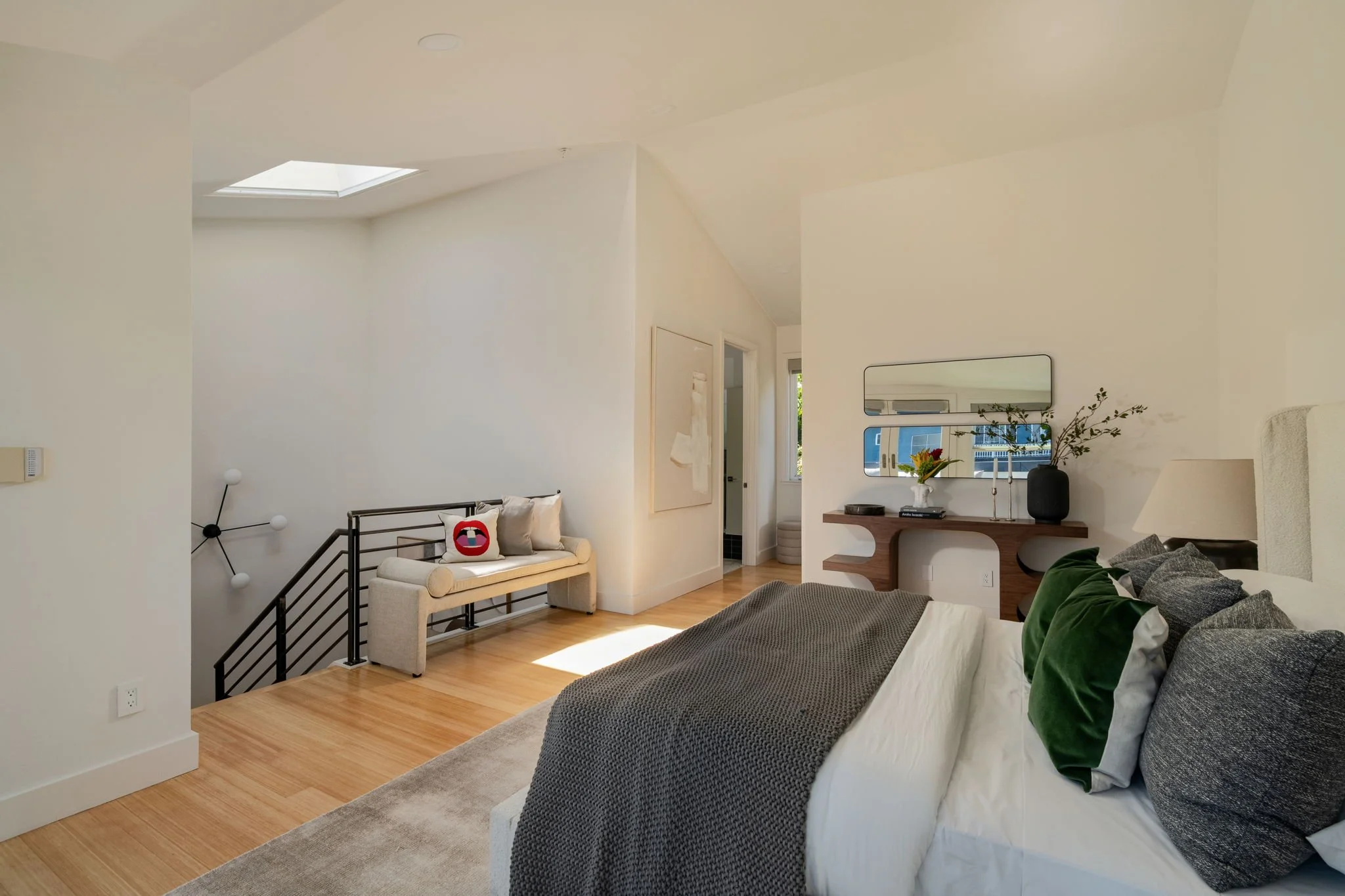 A modern bedroom with a skylight, white walls, and wood flooring. There is a beige armchair with a pink, black, and white pillow, and a bed with a gray blanket and various pillows. A wooden console with a black vase and decorative branches is against