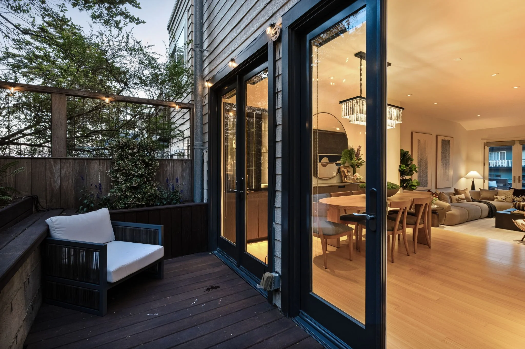 A cozy outdoor patio with wooden flooring and a sitting area with a white cushioned seat. It is connected to an indoor living room through large sliding glass doors, which reveal modern furniture and a chandelier inside. String lights are hung along 