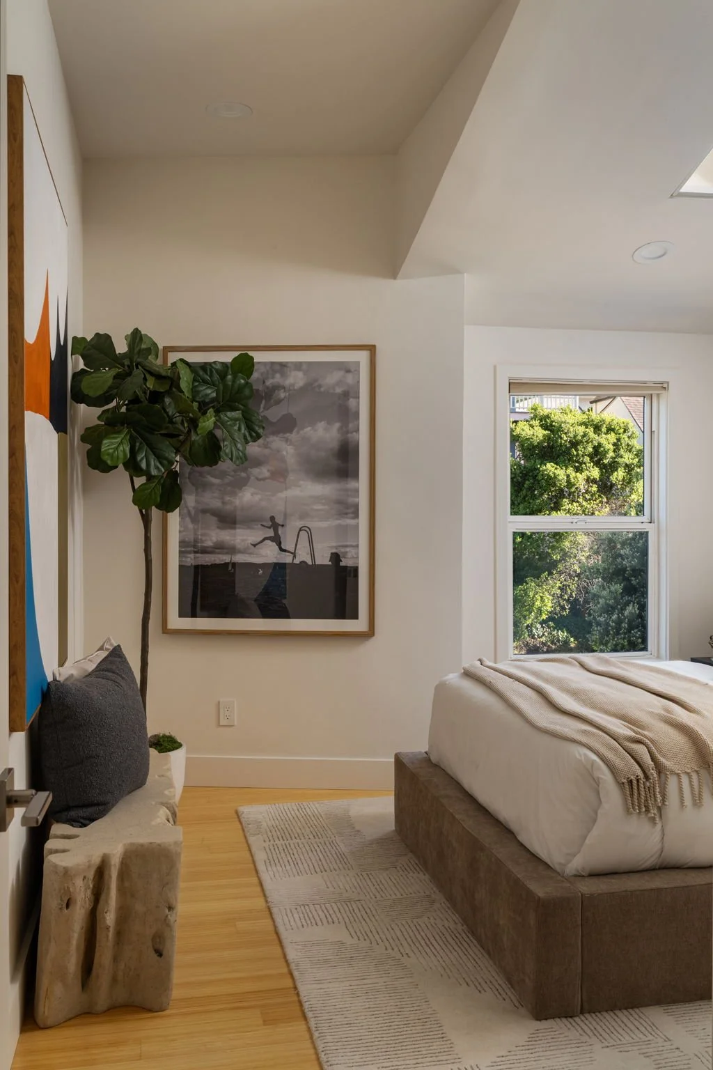 A bedroom with a large window showing green trees outside, a bed with beige blanket, a large framed black and white photograph of a person jumping into a pool, a tall green leafy plant in a white pot, and a gray cushion on a unique stone bench.
