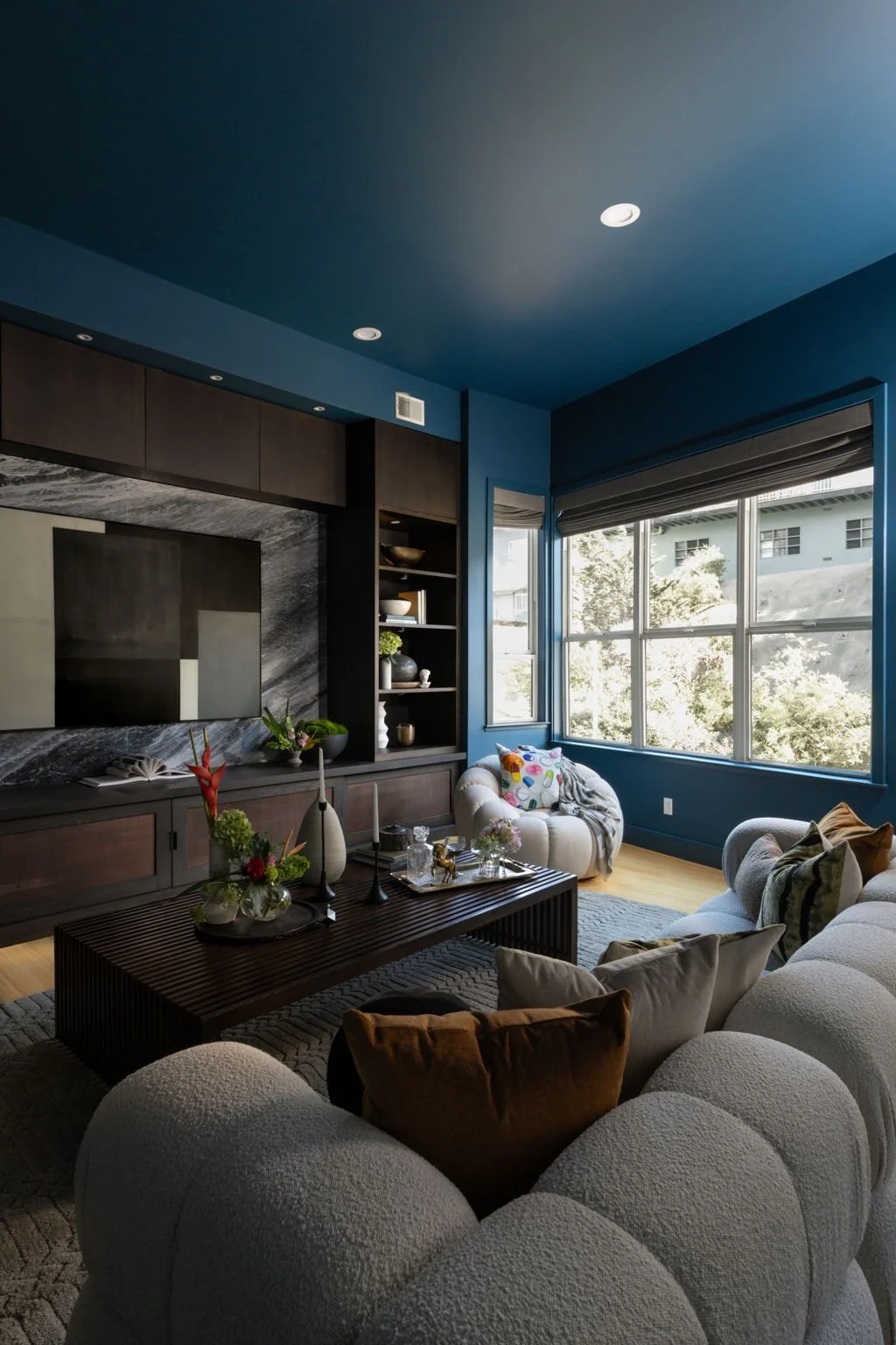 Modern living room with a large beige plush sofa, colorful pillows, a dark wooden coffee table with decorative items, and a large window with a view of trees and a building outside. The room has dark wood cabinetry and shelves on one wall, a blue cei