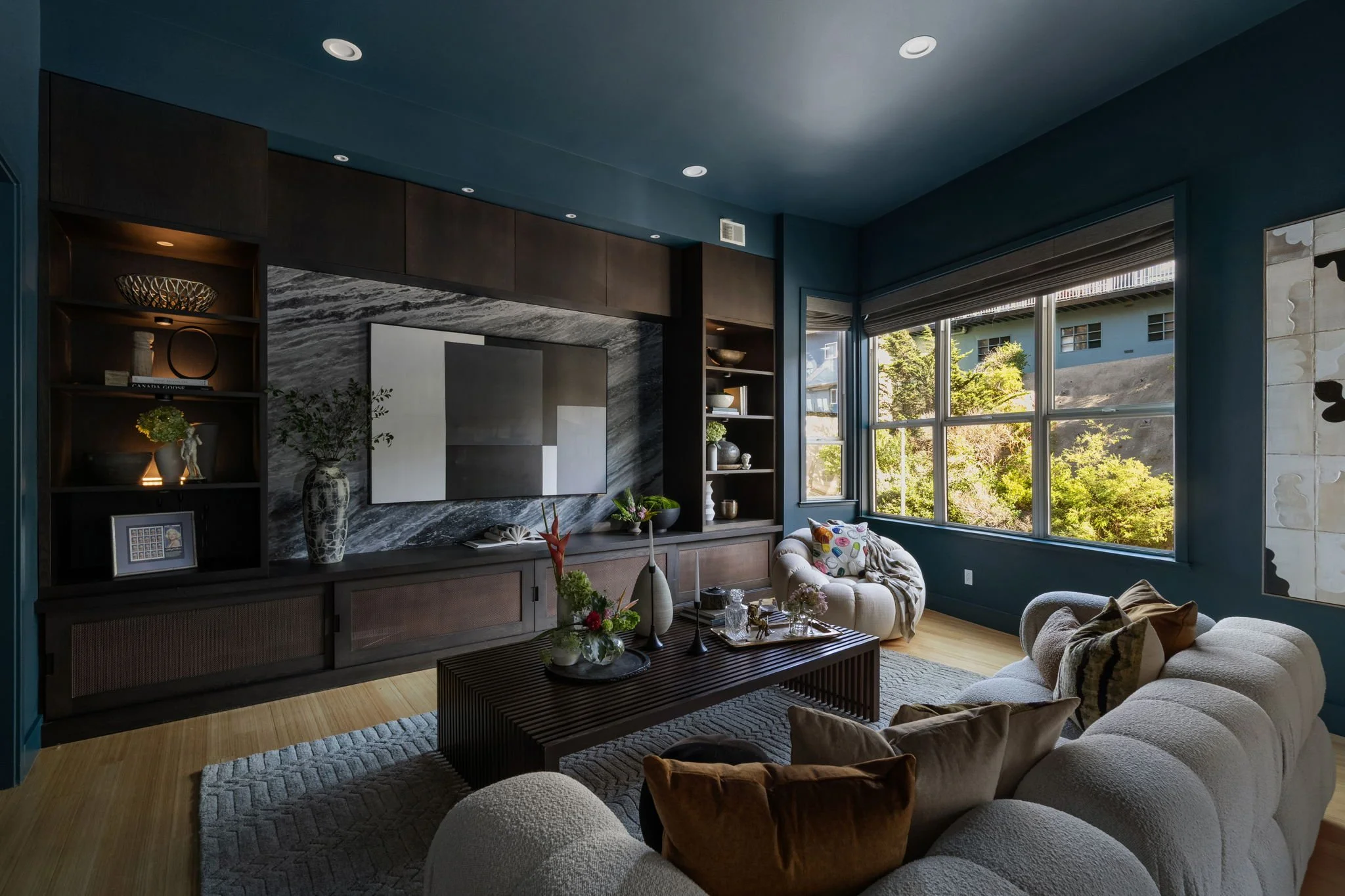 Living room with dark wood cabinetry, a large flat-screen TV, decorative shelves, and a beige sofa with colorful cushions. Large windows reveal a view of trees and neighboring buildings.