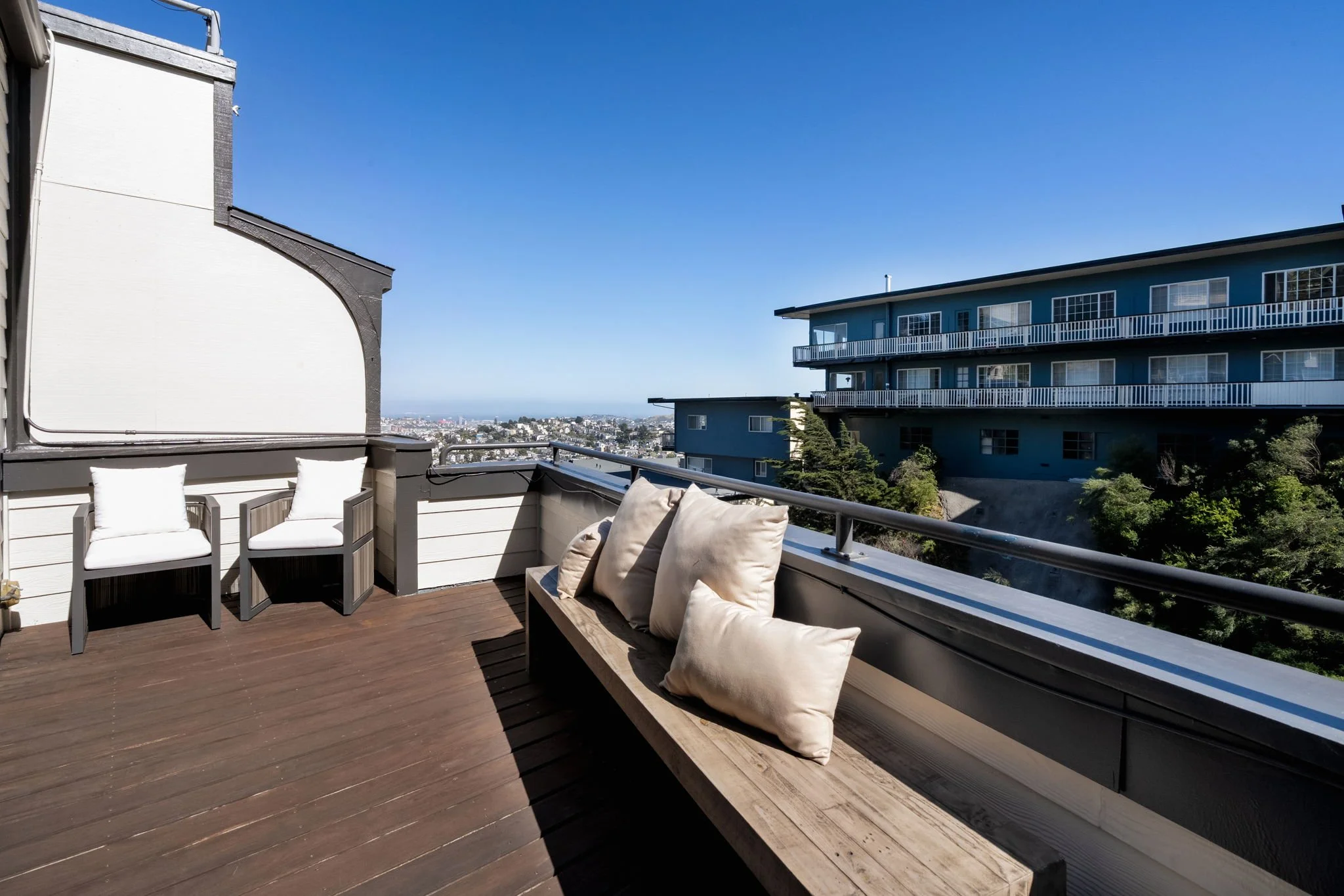 View of a rooftop deck with seating area, including chairs and cushions, overlooking a cityscape and a blue sky.