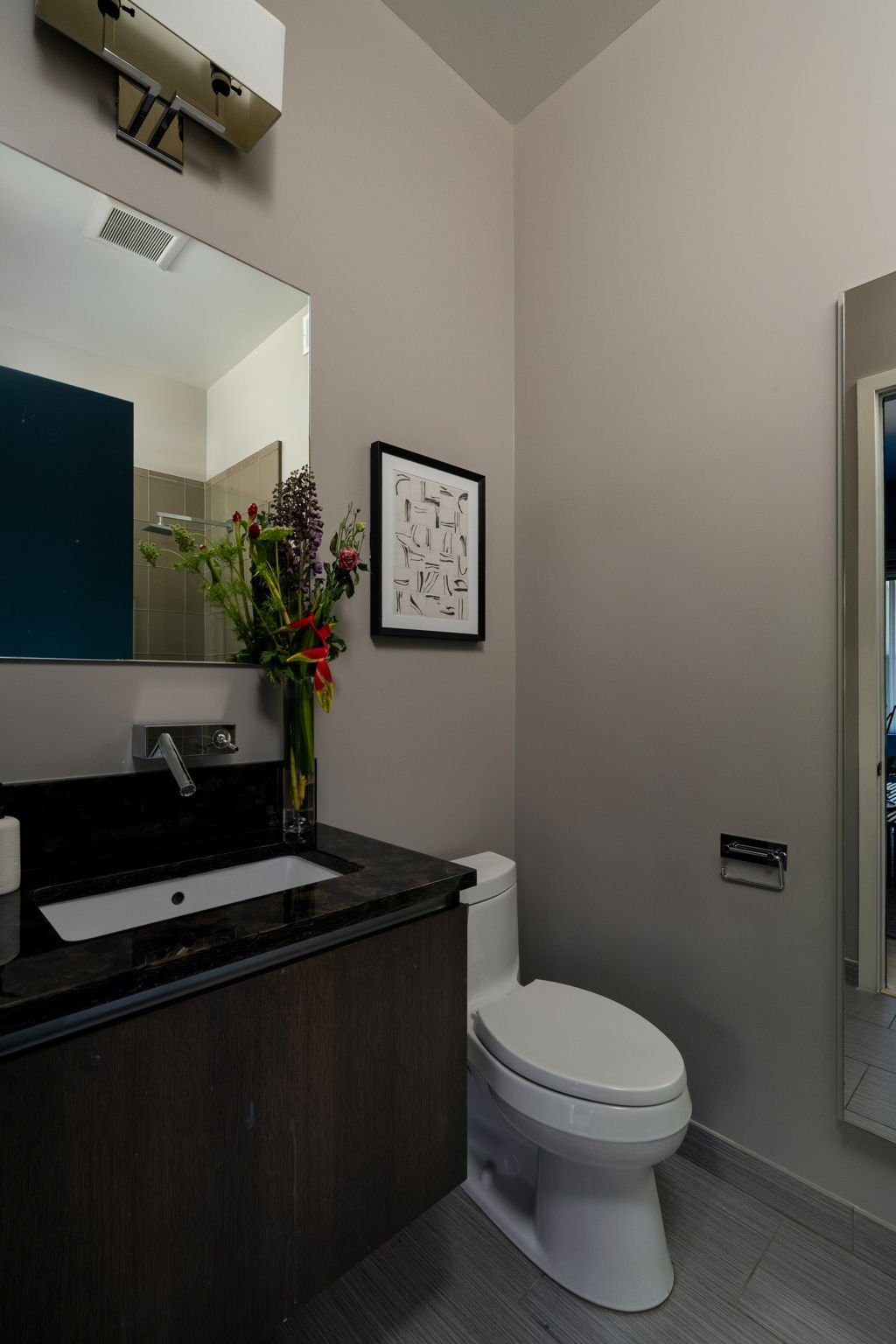 A modern bathroom with a white toilet, a black granite vanity with a sink, a large mirror, a flower vase, framed artwork, and a partial view of a shower area with beige tiles.