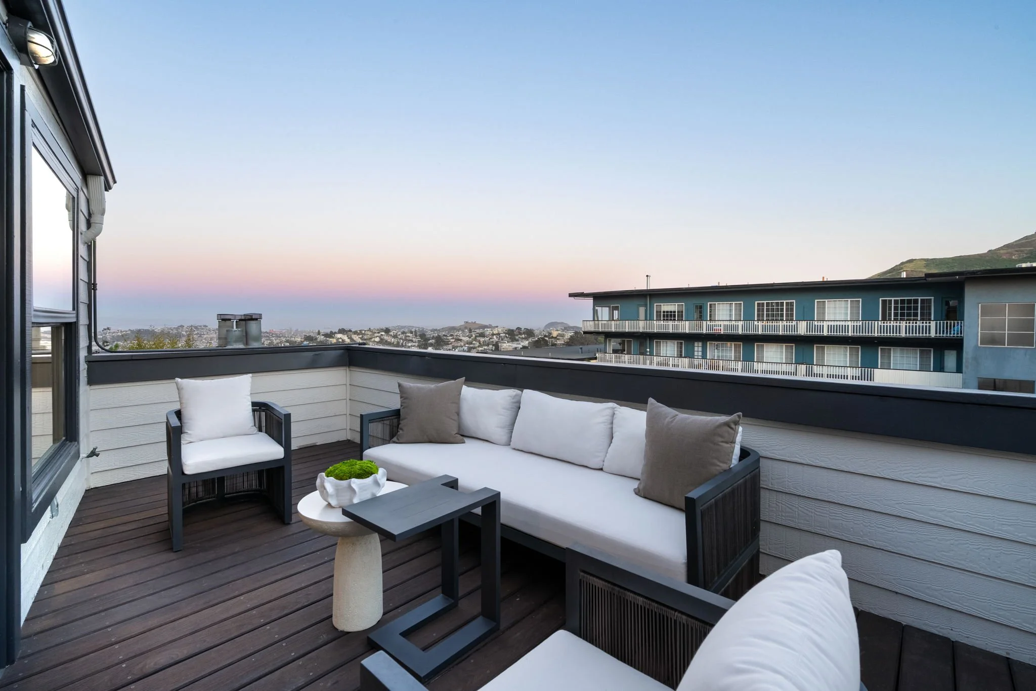 An outdoor balcony with a black railing, furnished with a black sofa with white and beige cushions, a black armchair with white cushions, a small black side table, and a beige stone pedestal table with a small green plant. The background shows neighb