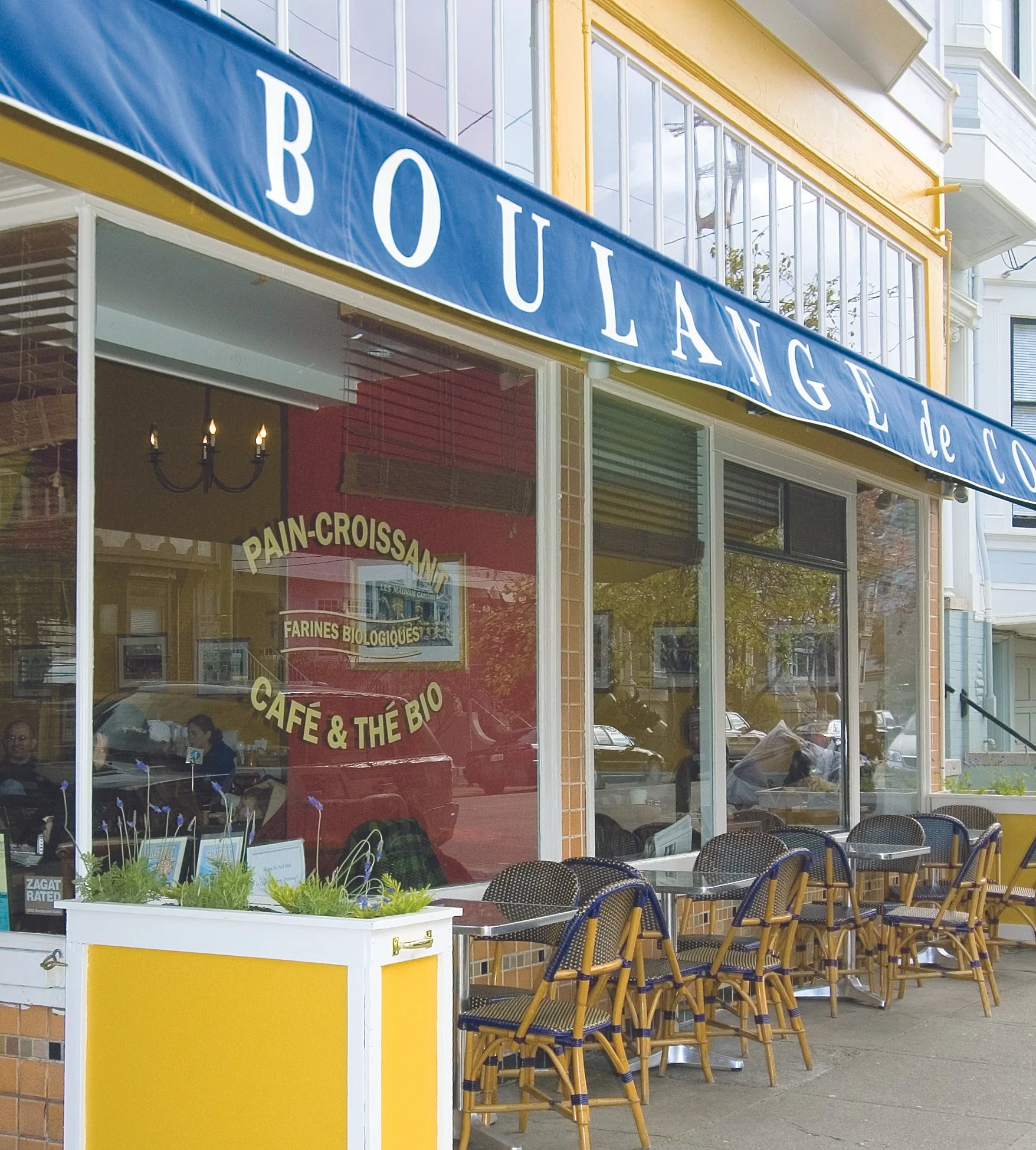 Outside view of a bakery called 'Boulangerie' with patio seating featuring wicker chairs with blue cushions, a yellow and white cabinet, and large windows reflecting nearby buildings.