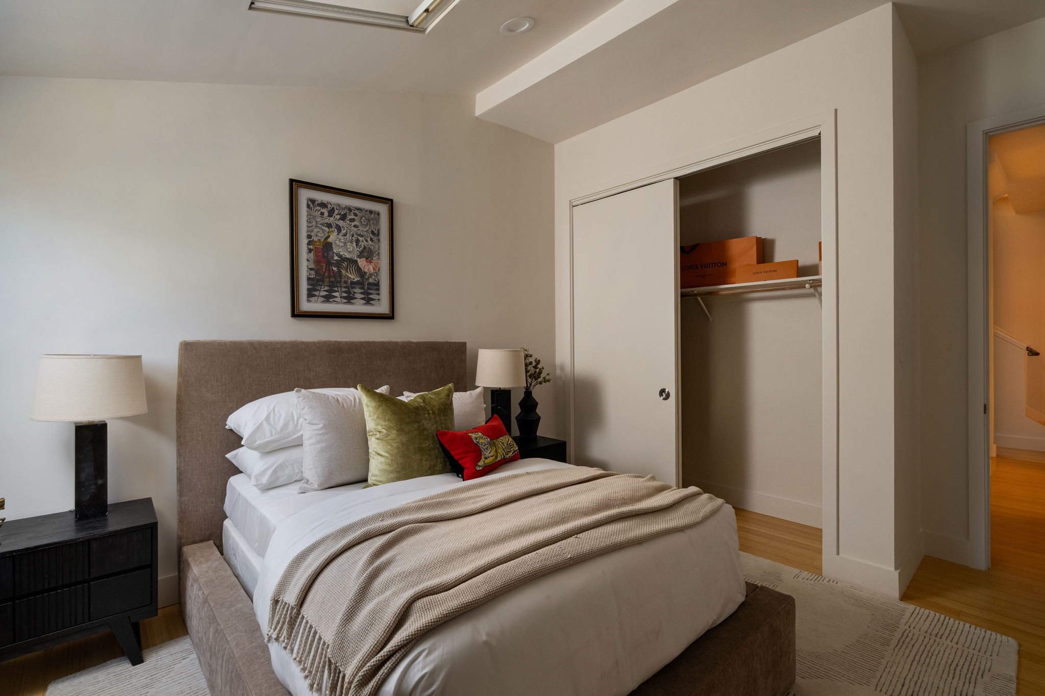 A bedroom with a bed featuring white bedding, a beige blanket, and decorative pillows, including one with a tiger design. There are two black nightstands with white lamps, a framed artwork on the white wall, and a closet with sliding doors, one parti