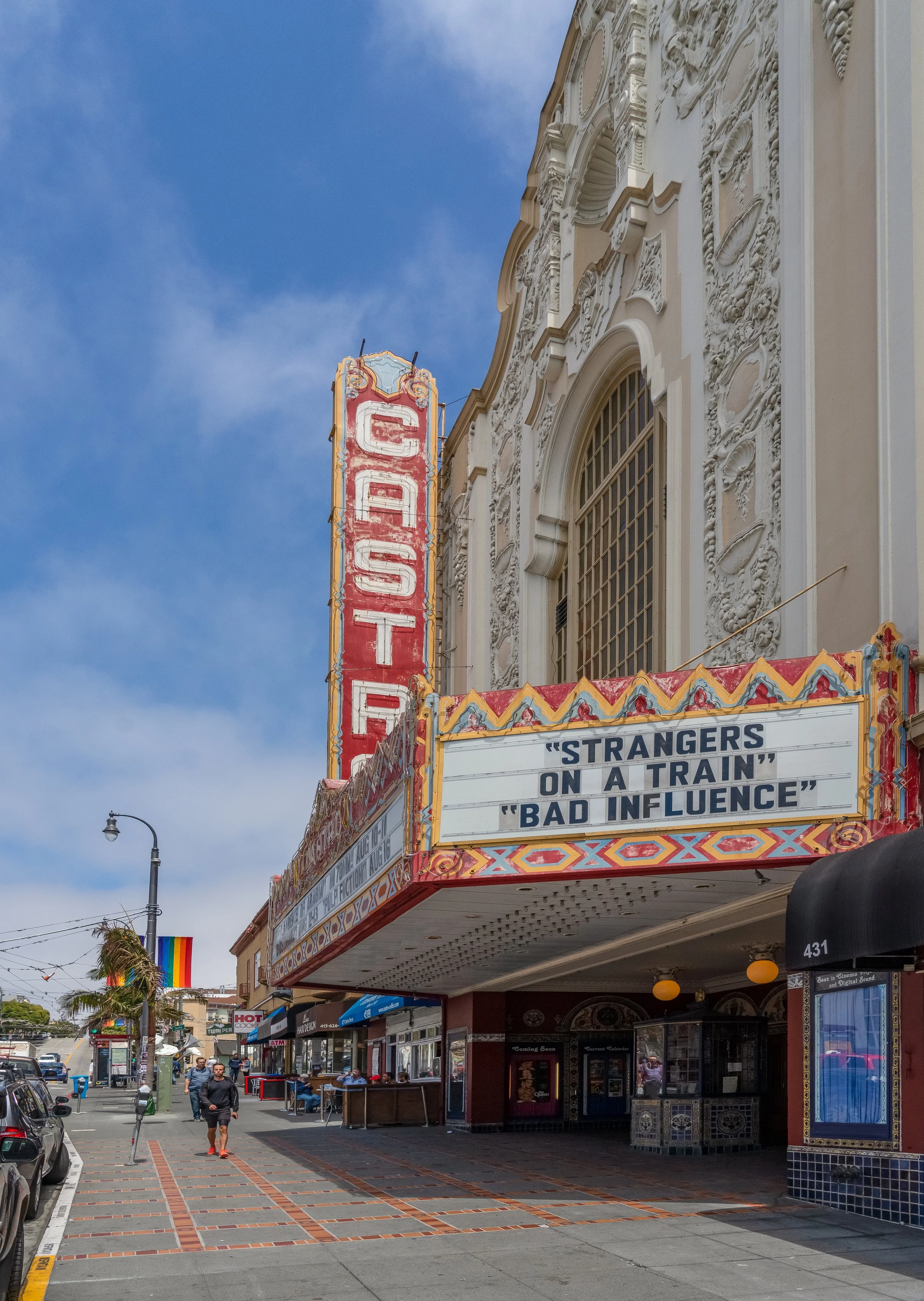 Street view outside a theater with a marquee displaying quotes from movies, people walking, a rainbow pride flag, and building facades with ornate architecture.