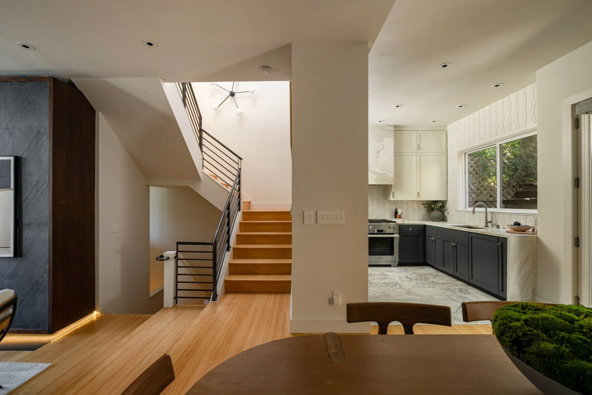 Interior view of a modern home showing a staircase with wooden steps and black metal railing, a kitchen with white upper cabinets, black lower cabinets, marble countertops, and a window with outdoor greenery visible.