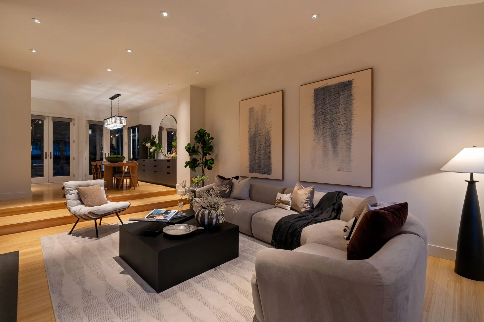 A modern living room with a beige sofa, a black coffee table with decor, a white armchair, and a living plant. There are three large framed abstract art pieces on the wall, a lamp, and a collection of pillows and throws on the sofa. The room is well-