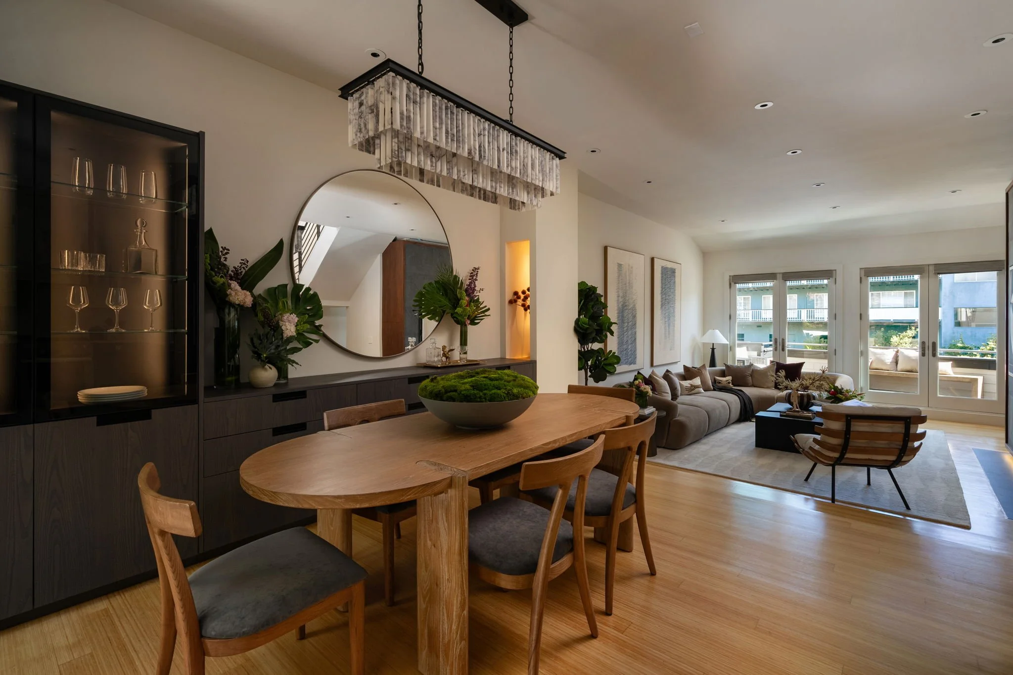 A modern living room with large windows, a beige sectional sofa with multiple cushions, a wooden coffee table, and a wooden rocking chair. There is a large round mirror on the wall, framed artwork, potted plants, and a chandelier hanging from the cei