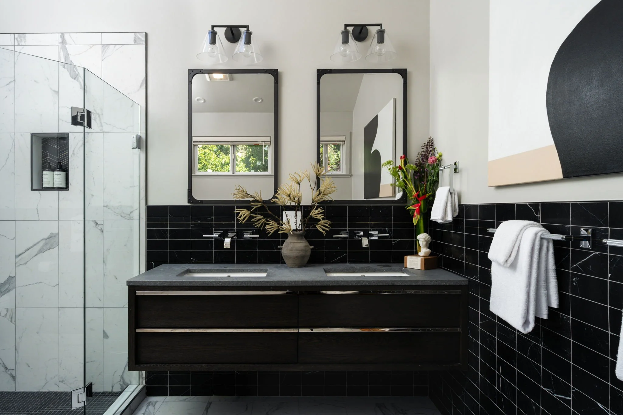 A modern bathroom with black and white tile decor. It features a dual sink vanity with a vase of flowers, large mirrors, a black framed artwork, a glass shower enclosure, and white towels.