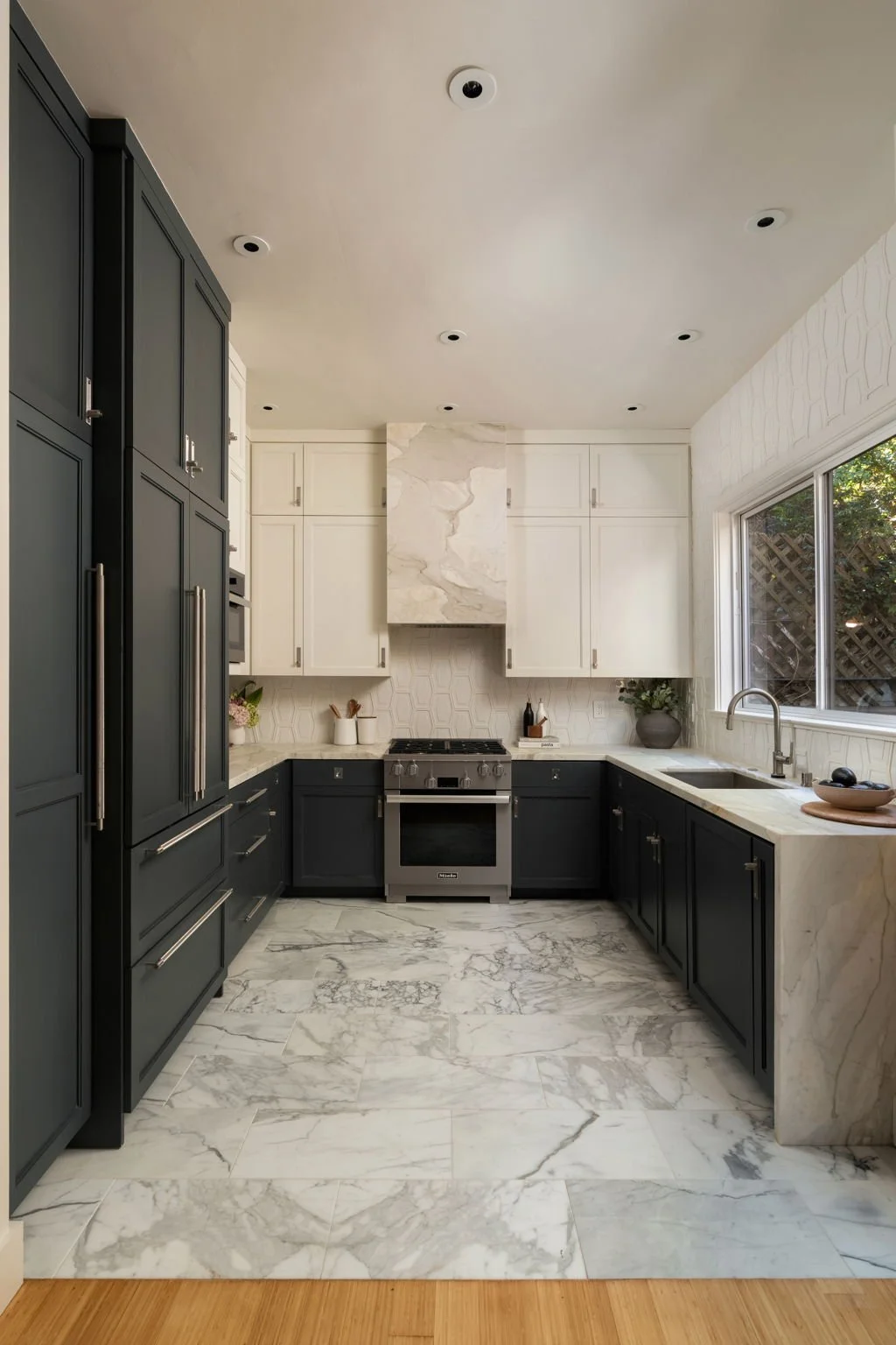 Modern kitchen with black and white cabinetry, marble countertops and backsplash, stainless steel stove, and large window showing outdoor greenery.