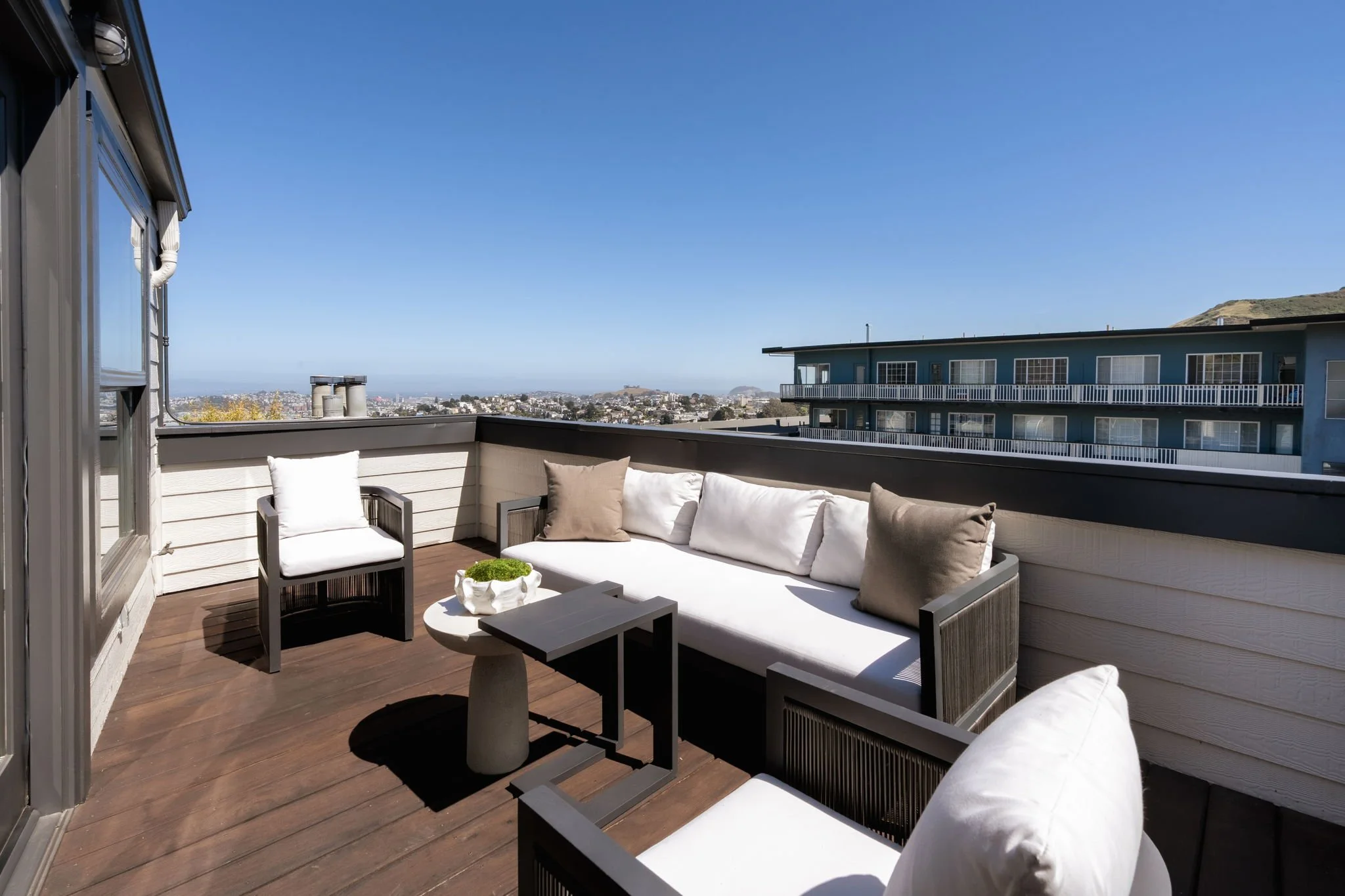 Outdoor balcony with white cushioned seating, a small table with a bowl of green plants, and a view of neighboring buildings and hills under a clear blue sky.