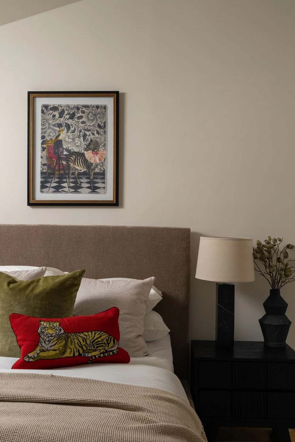 A close-up of a bed with multiple pillows, including a red pillow with a tiger illustration, a beige pillow, and a white pillow. To the right, there is a black nightstand with a white lamp and a decorative plant. A framed artwork featuring a zebra, a