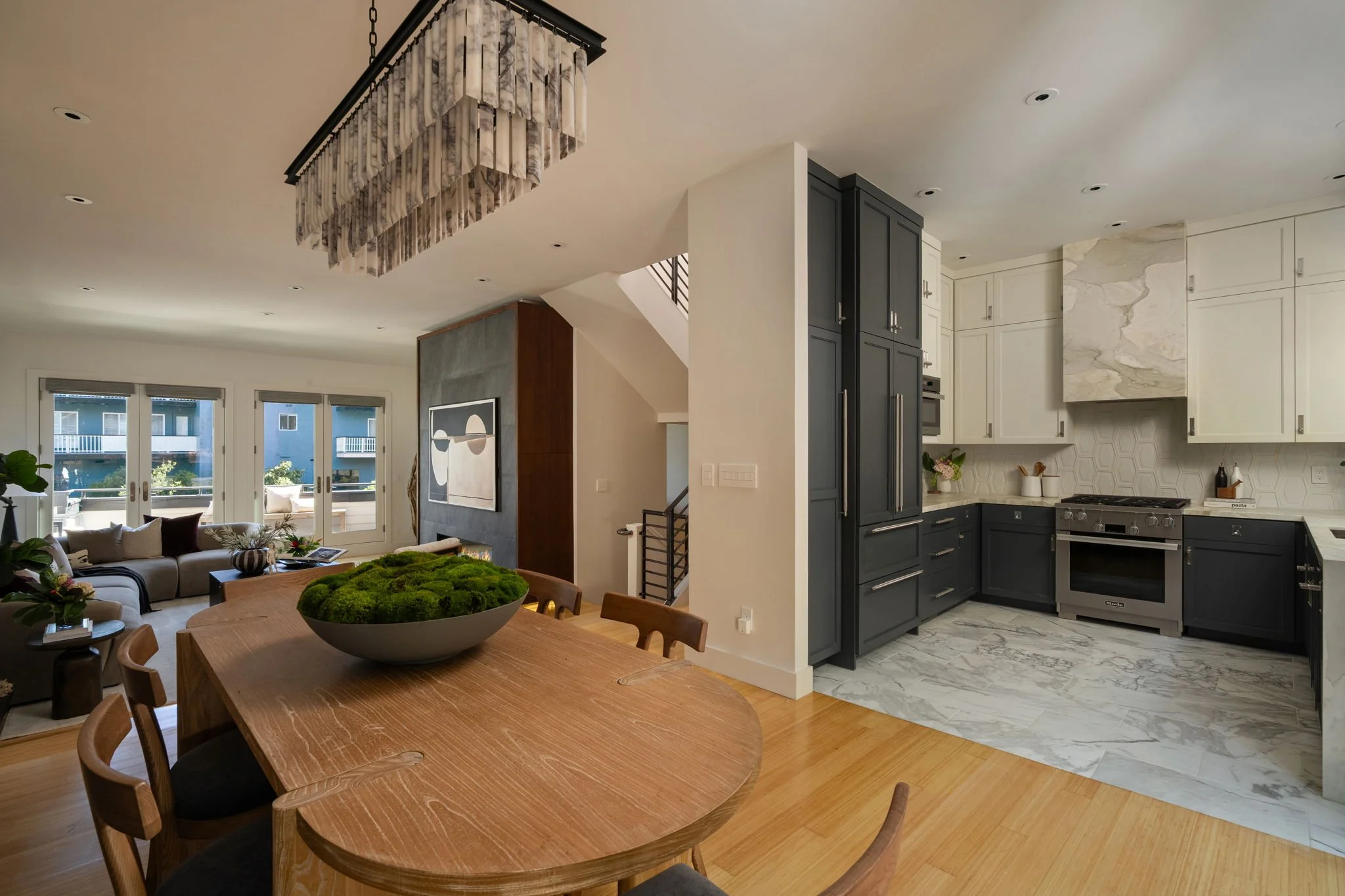Open-concept living room and kitchen with modern design, featuring a wooden dining table, a chandelier, beige sofa, large windows, and navy blue kitchen cabinets with marble countertops.