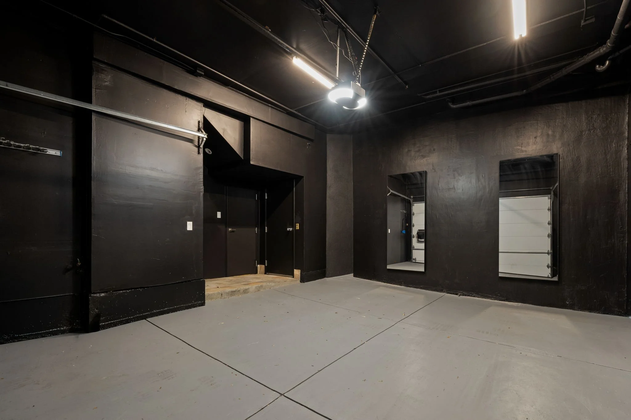 Interior view of a dark basement or garage with black painted walls and ceiling, concrete floor, and fluorescent lighting. There are two mirrors on the right wall, a door on the left wall, and some hanging chains or wires.