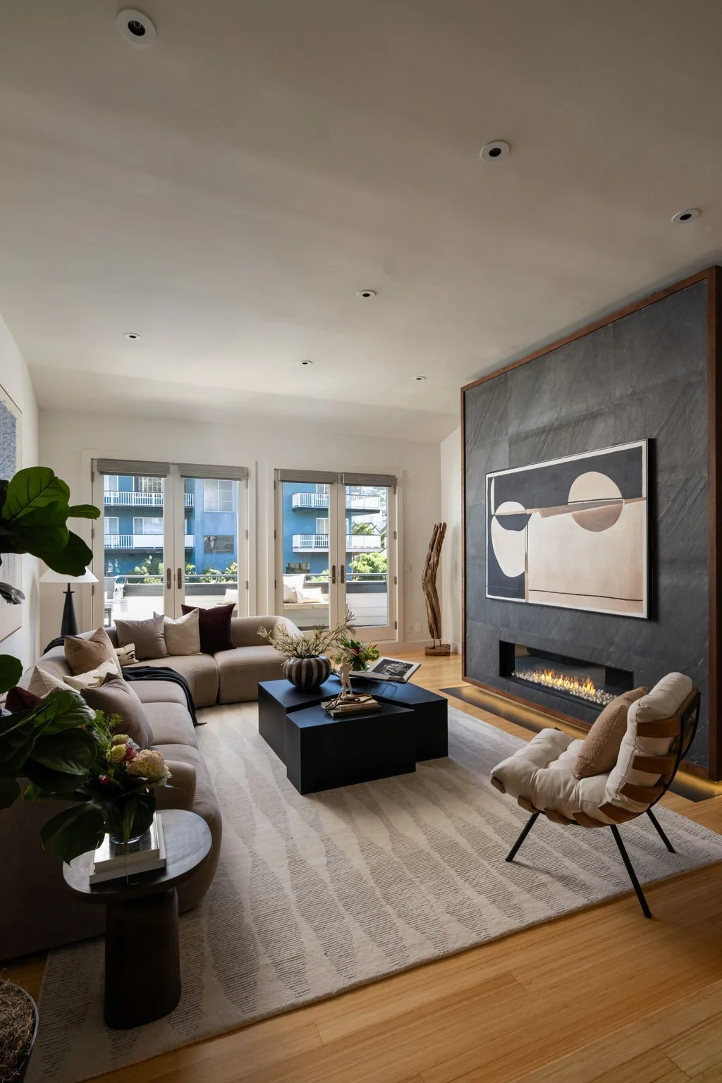 Modern living room with beige sectional sofa, black coffee table, abstract artwork above fireplace, and large windows with views of balconies outside.