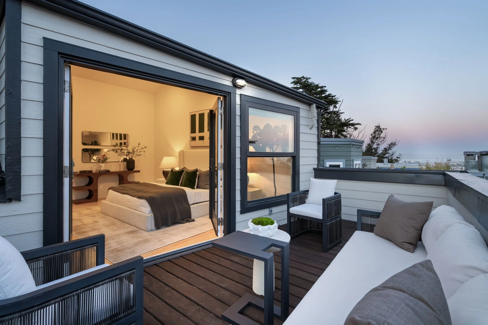 Outdoor rooftop patio with seating, overlooking trees and city skyline at sunset, adjacent to a modern bedroom with open sliding door.