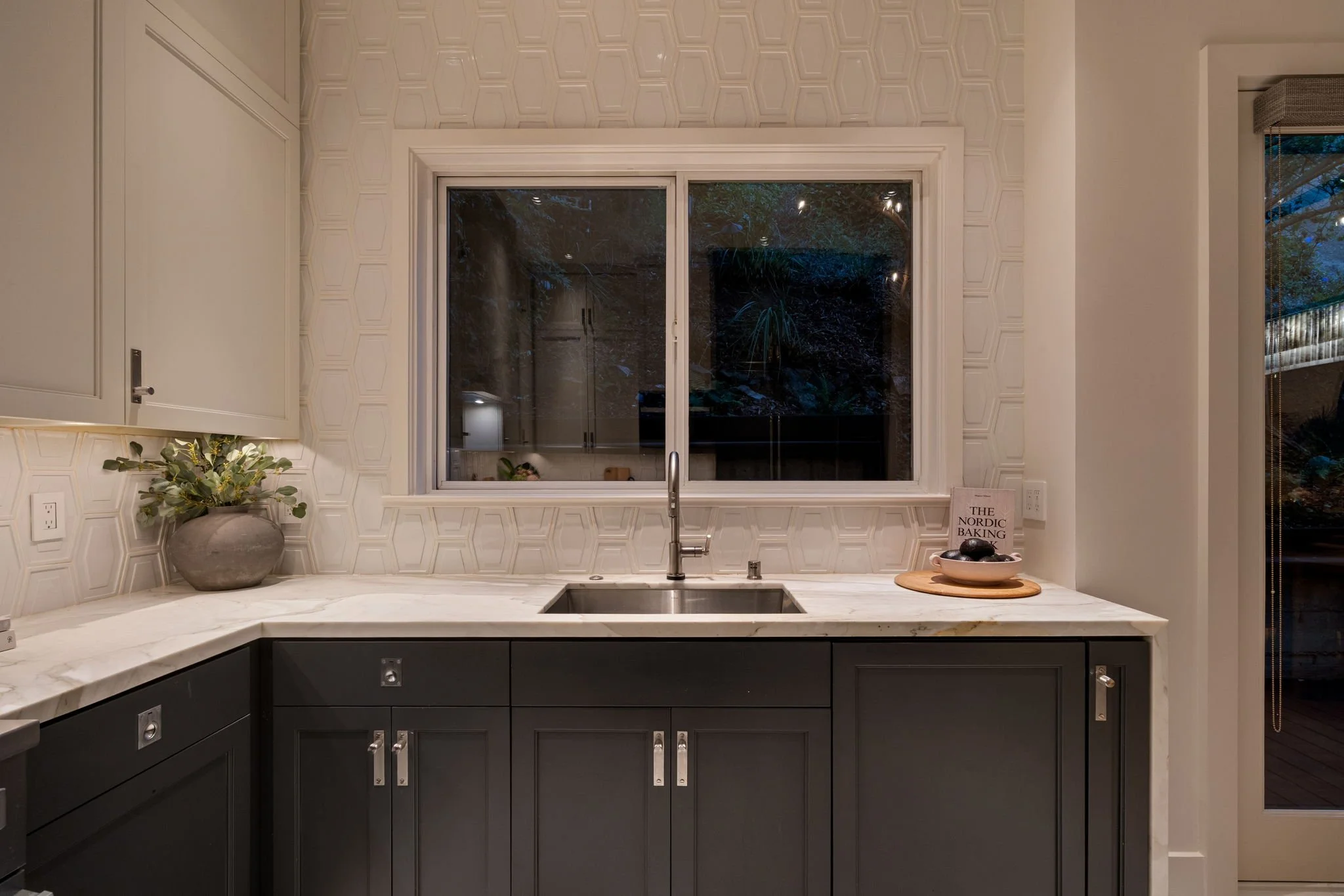 Modern kitchen with gray cabinets, white marble countertop, and a large window over the sink. There is a potted plant and a cookbook on the counter.