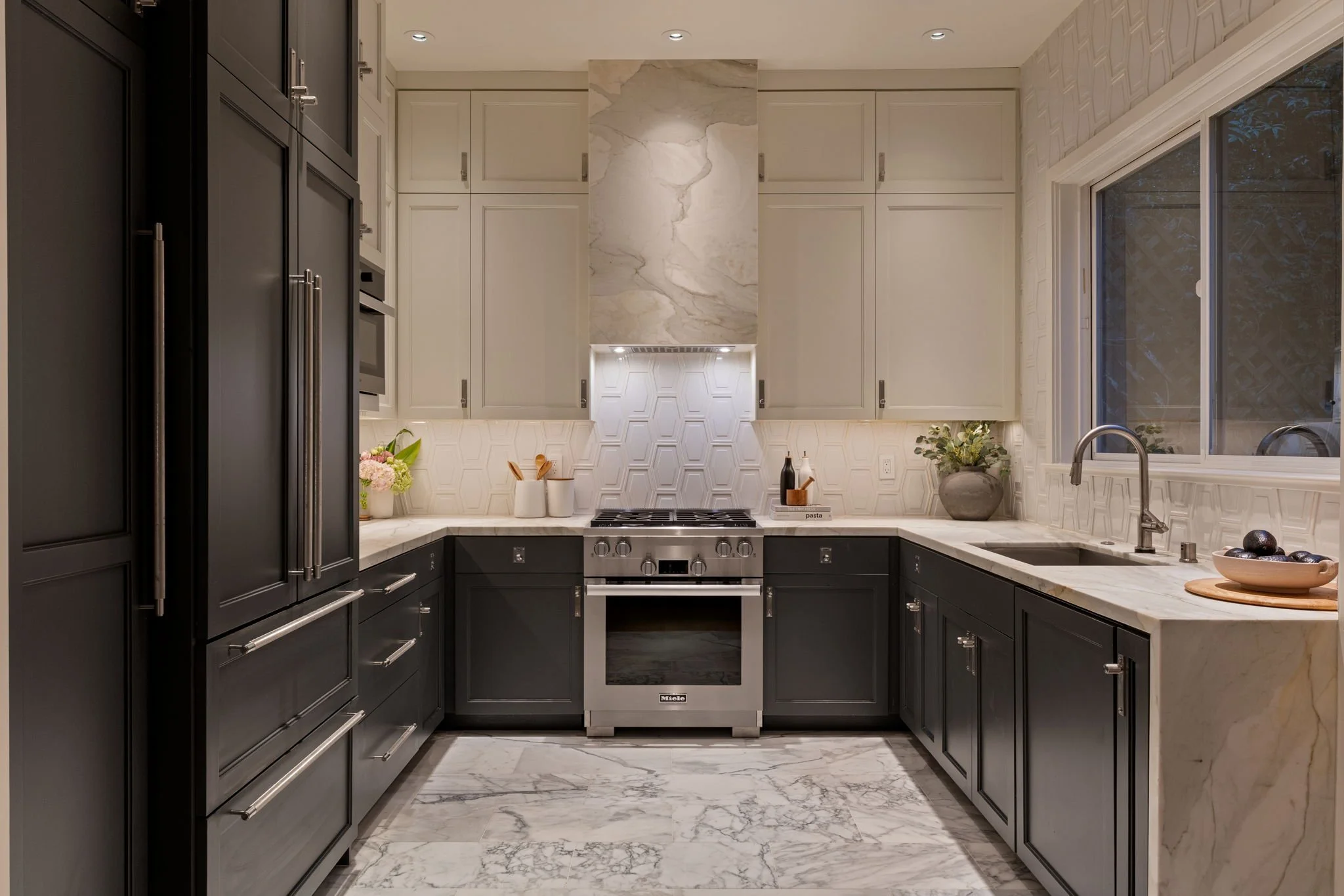 Modern kitchen with dark lower cabinets, white upper cabinets, marble backsplash, and stainless steel appliances. There is a window above the sink, potted plants, and kitchen utensils on the counter.