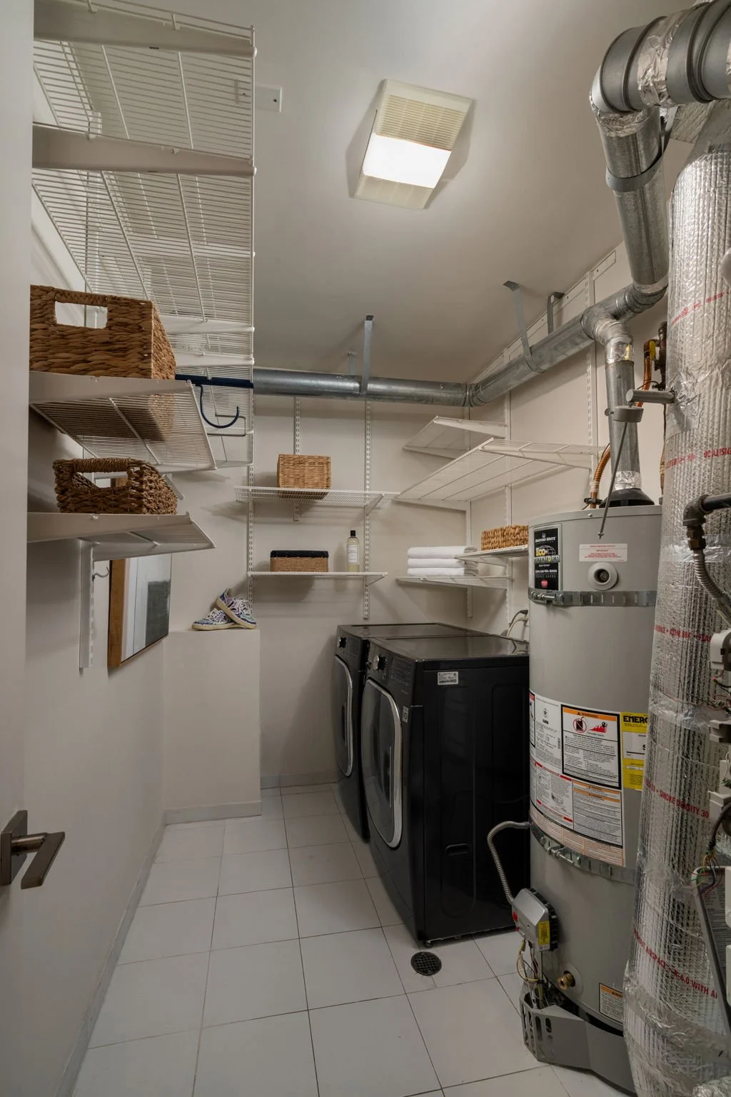 Laundry room with white walls and tiled floor, featuring front-loading washer and dryer, wire shelves with baskets, a water heater, and utility pipes.