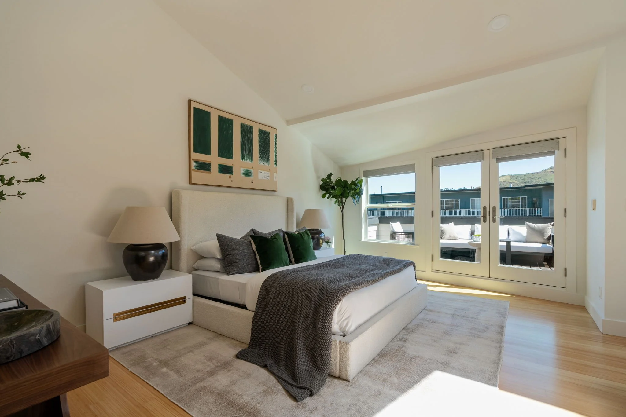 A bedroom with a white bed, dark pillows, and a gray blanket near a large window. There are two nightstands with lamps, a piece of modern art on the wall, a houseplant, and a balcony with outdoor seating