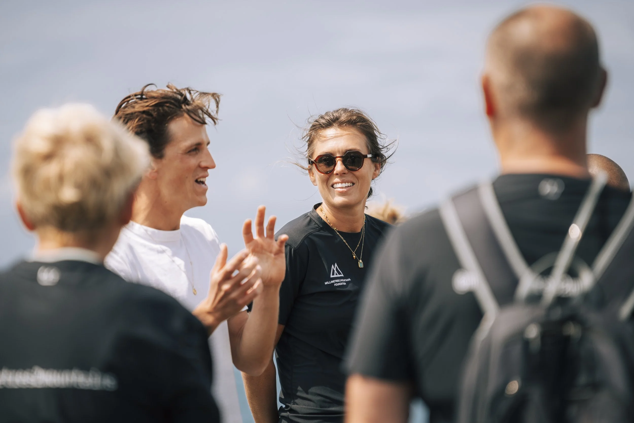 Group of people outdoors, engaged in conversation, with one woman wearing sunglasses smiling amid a bright sky.