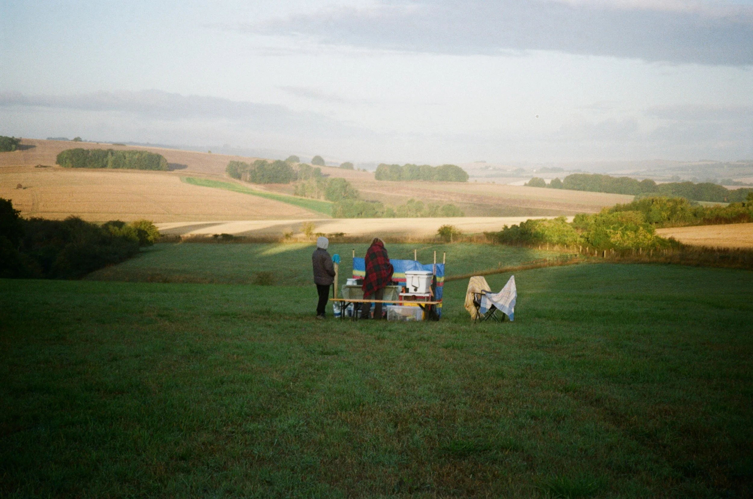 Two people standing on a grassy field near a table with ice coolers and chairs, overlooking a countryside landscape with rolling hills and fields under a cloudy sky.