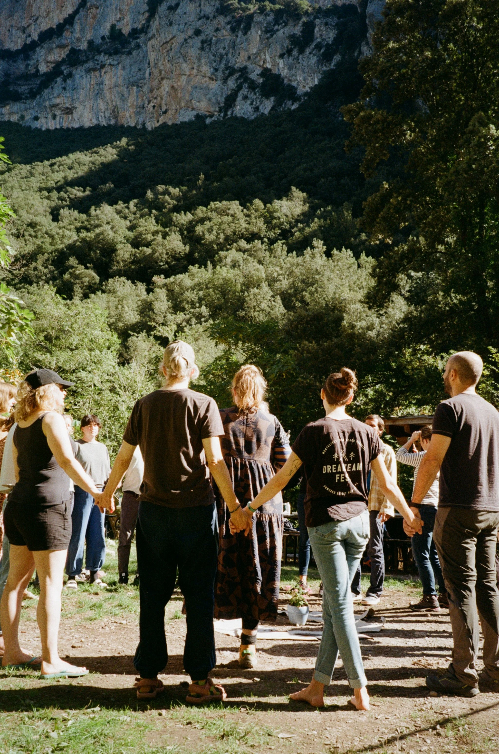 Group of people holding hands in a circle outdoors with trees and rocky hillside in the background.