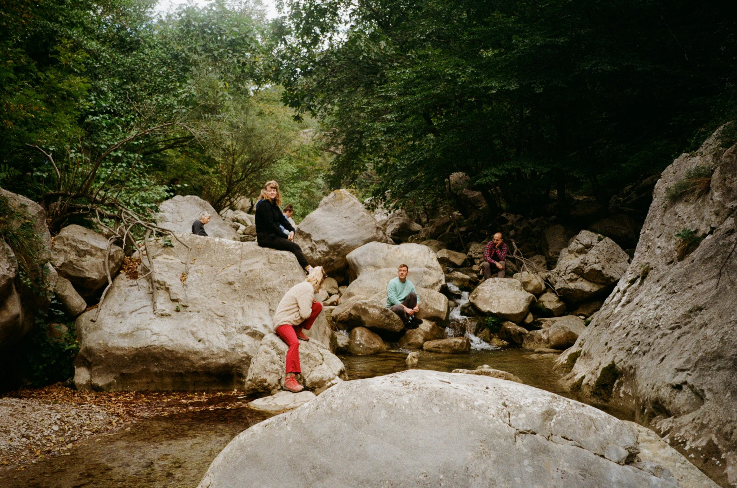 People sitting and standing on large rocks by a river in a forested area.