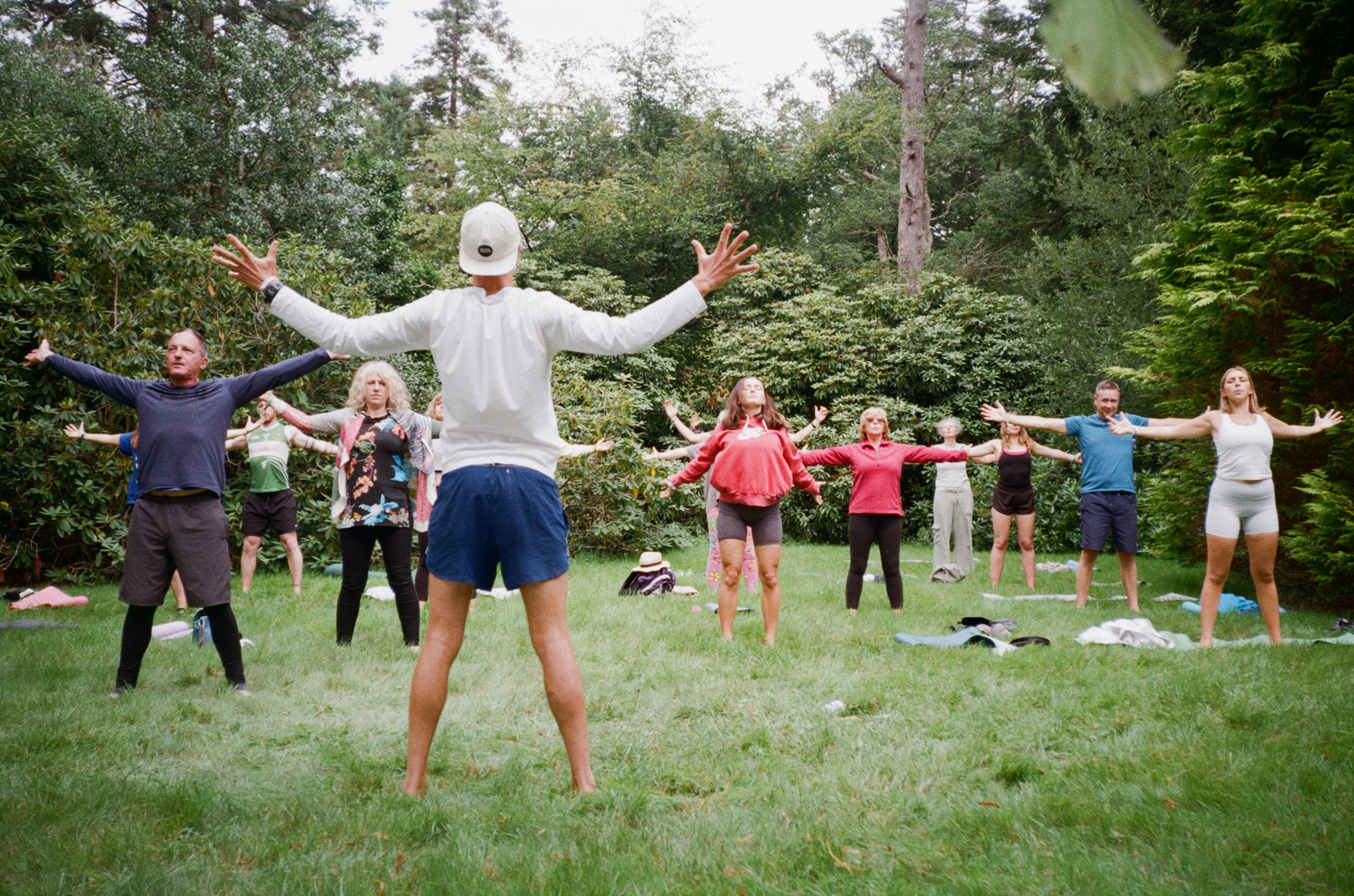 Group of people practicing yoga outdoors in a forested area, standing with arms outstretched.
