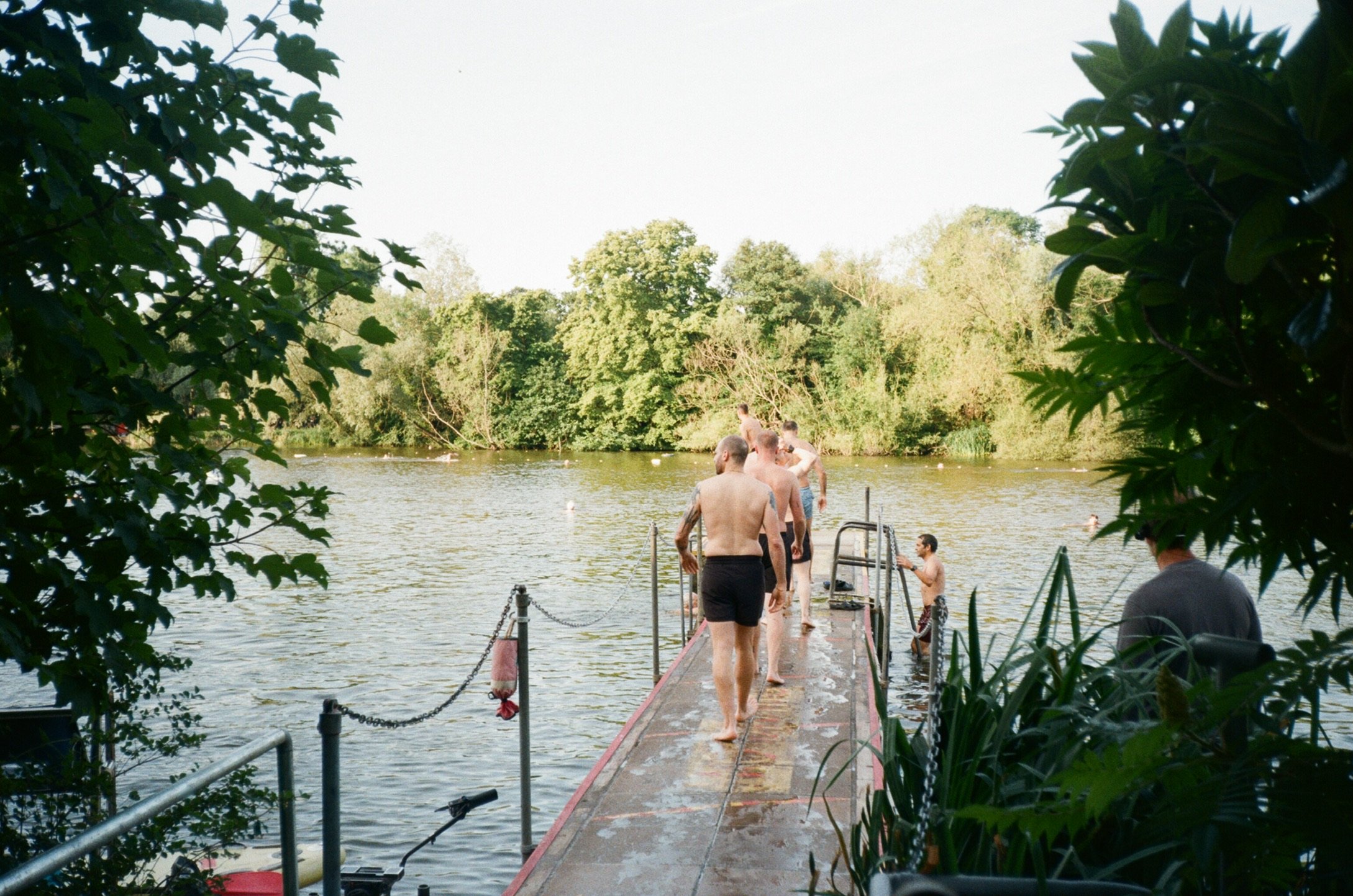 People in swimsuits walking on a dock by a lake, surrounded by lush trees, with some ducks swimming in the water.