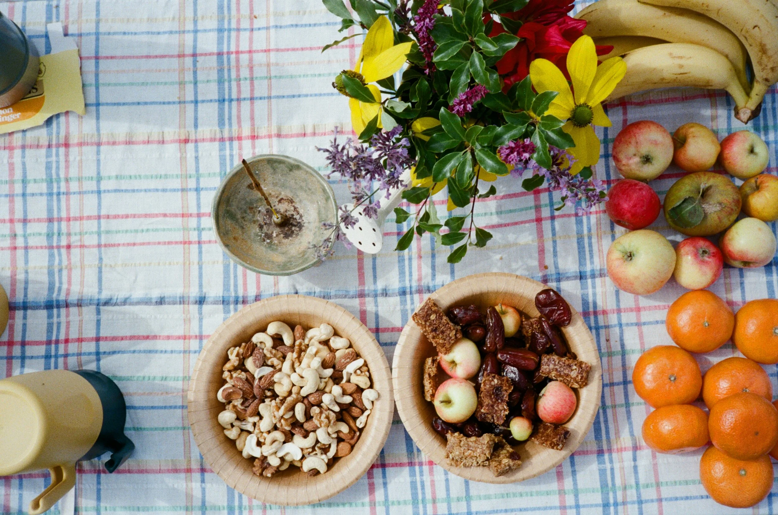 A table with a checkered tablecloth holding a colorful array of fruits, nuts, and snacks, including apples, bananas, oranges, a bowl of mixed nuts, and other snacks, along with a small flower arrangement.