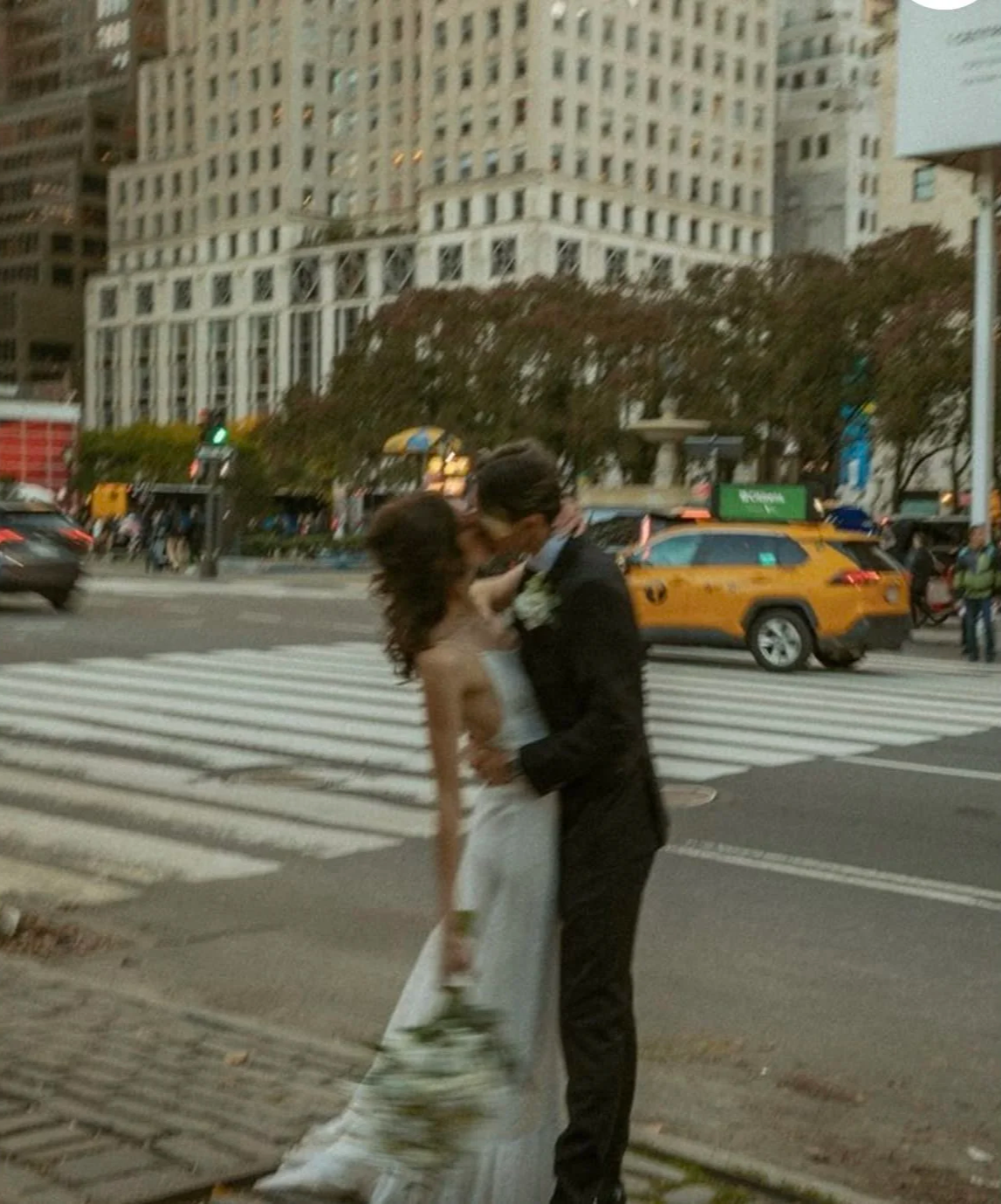 A couple kissing on a city crosswalk with buildings, yellow taxis, and pedestrians in the background.