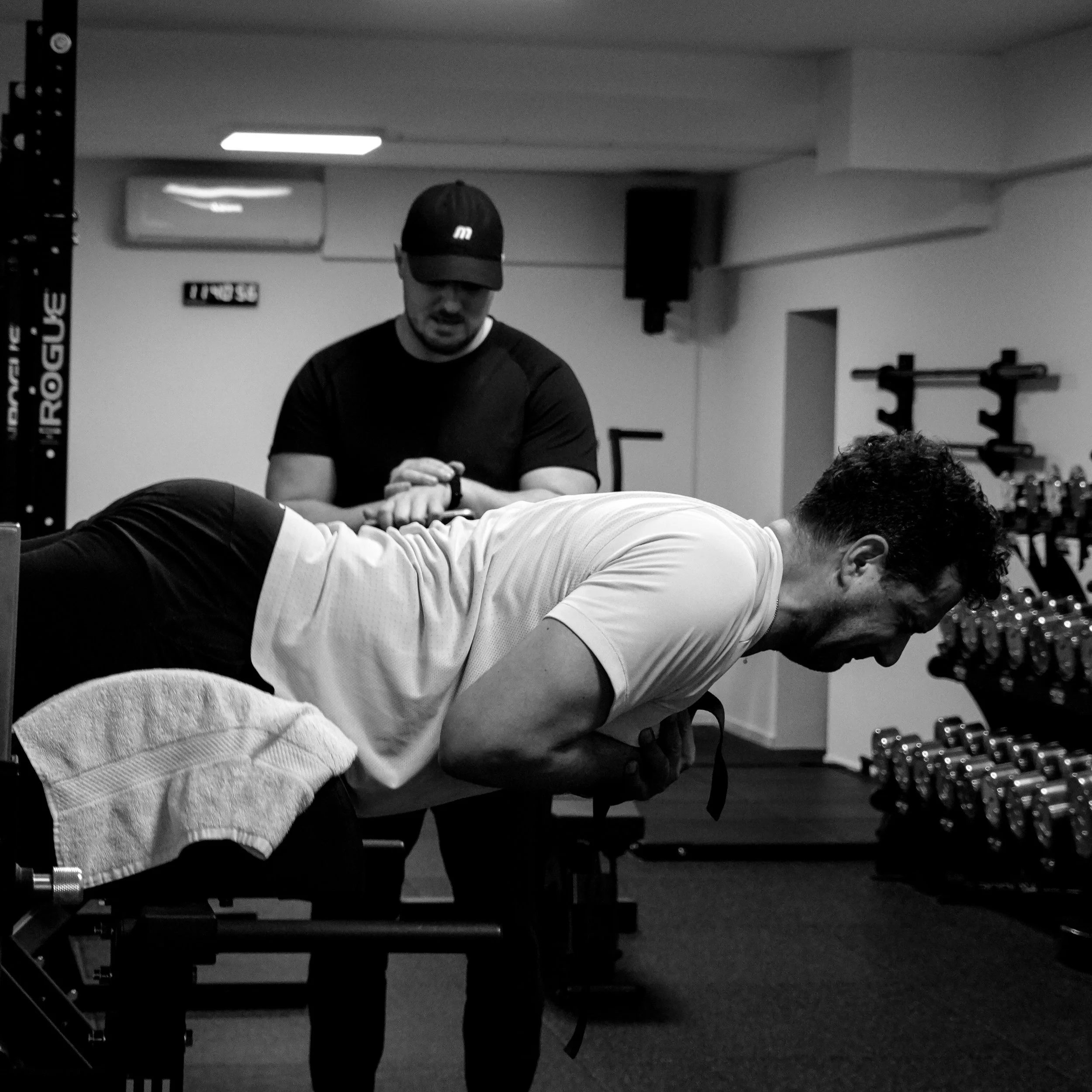 A man in a white athletic shirt is performing a plank exercise on a padded exercise bench in a gym. In the background, another man in a black cap and shirt is assisting or coaching. The gym has weightlifting equipment and a mirror on the wall.